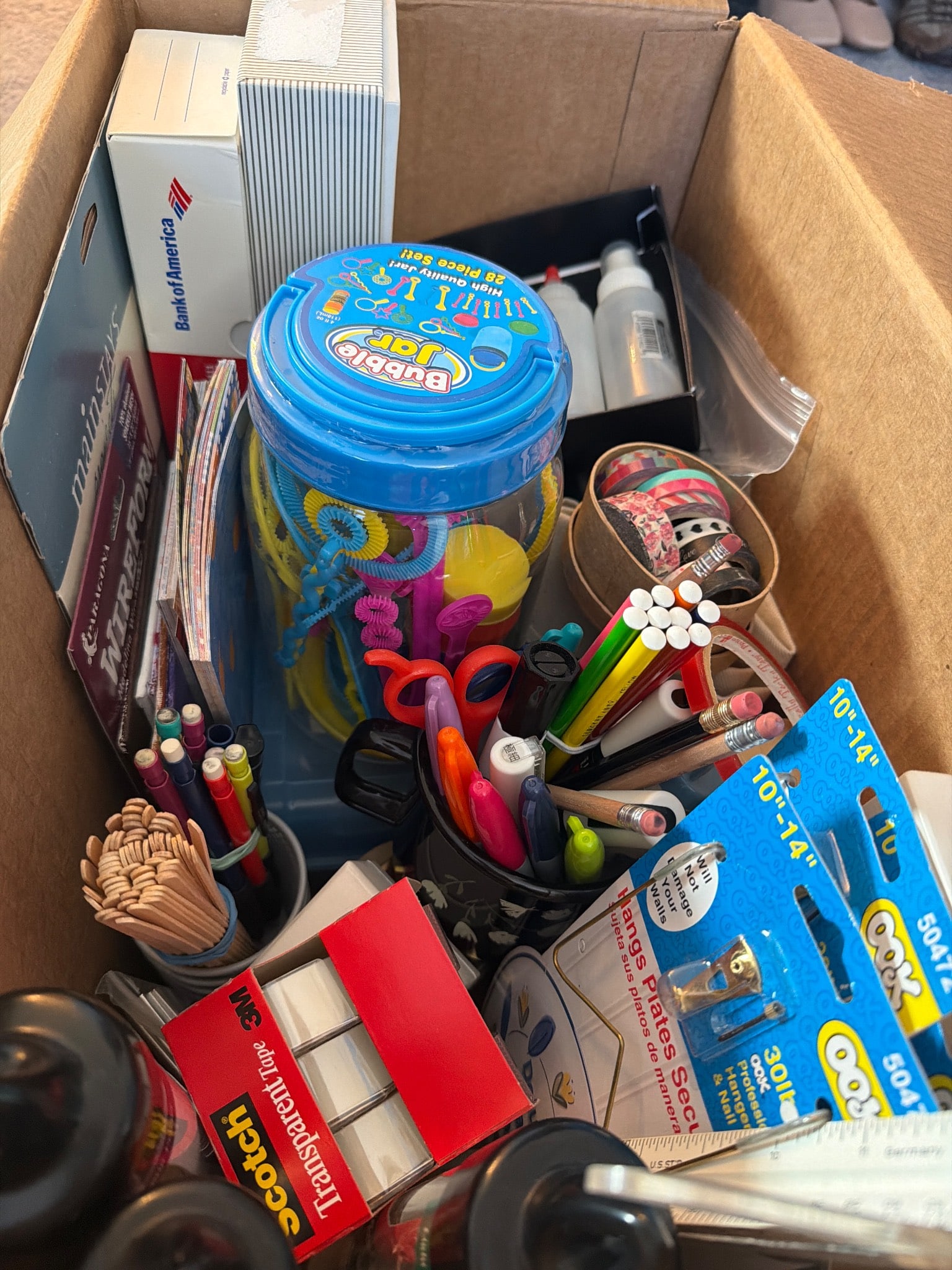Craft and Office Supplies Including Washi Tape and Beads: A box containing an assortment of craft and office supplies, including washi tape rolls, scissors, colored pencils, pens, markers, straws, beads in a labeled container, a jar of bubble wands, metallic