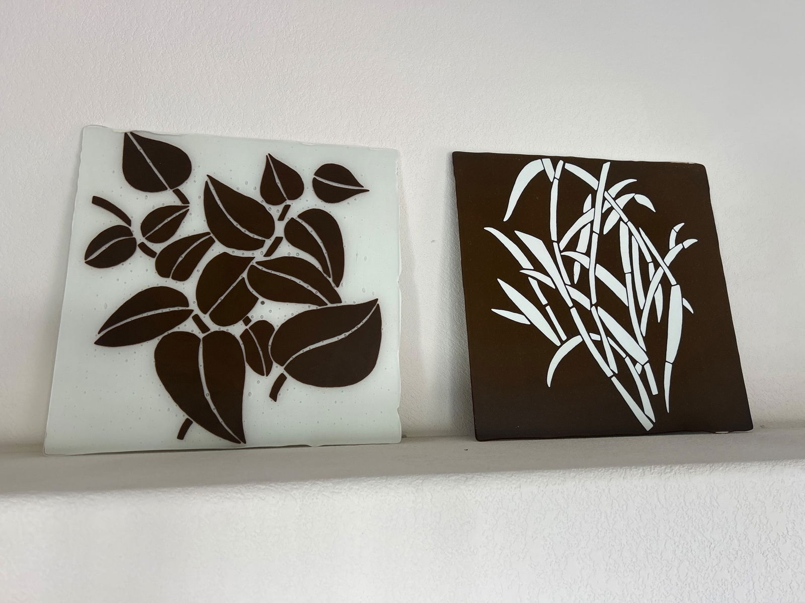 Two Decorative Ceramic Tiles with Botanical Designs: Two ceramic tiles featuring botanical designs. One tile showcases brown foliage on a white background, while the other highlights white foliage on a brown background. Both have a glossy finish and sha