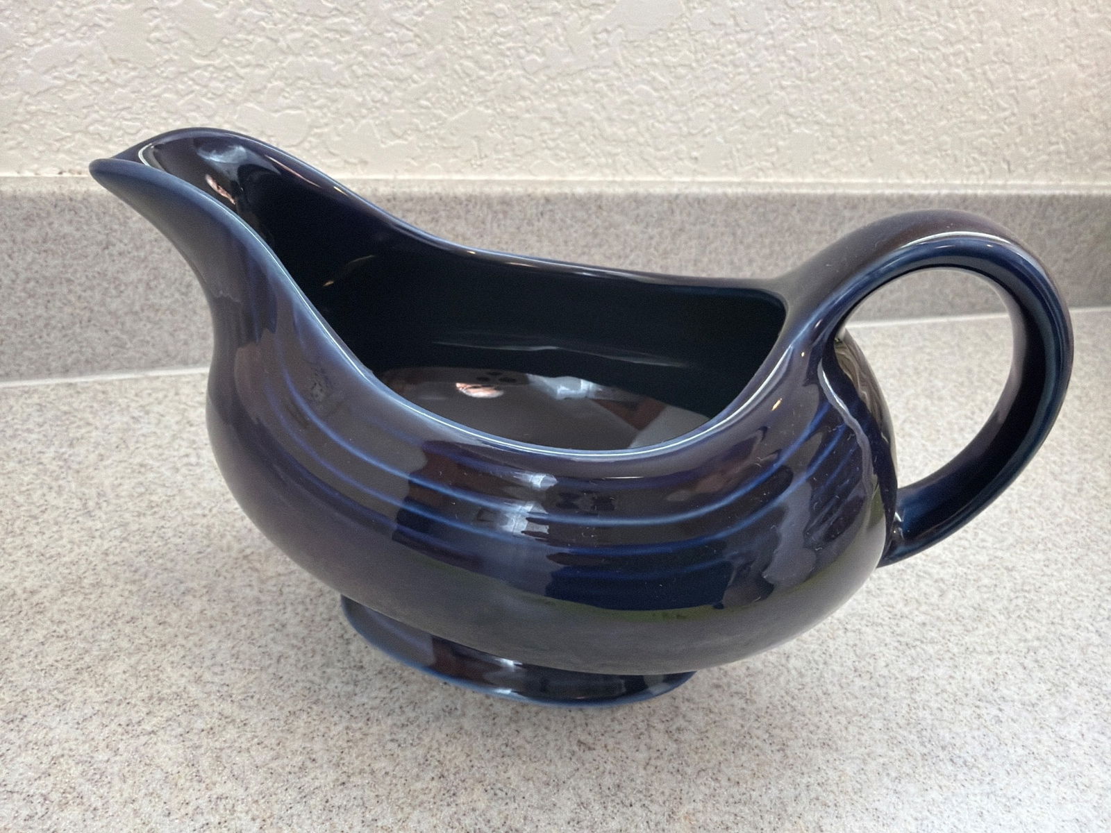 Vintage Blue Ceramic Gravy Boat: Vintage blue ceramic gravy boat featuring a curved handle and spout design. The glossy finish and subtle ridges on the body add charm. The base shows visible wear consistent with age and use. Perfect