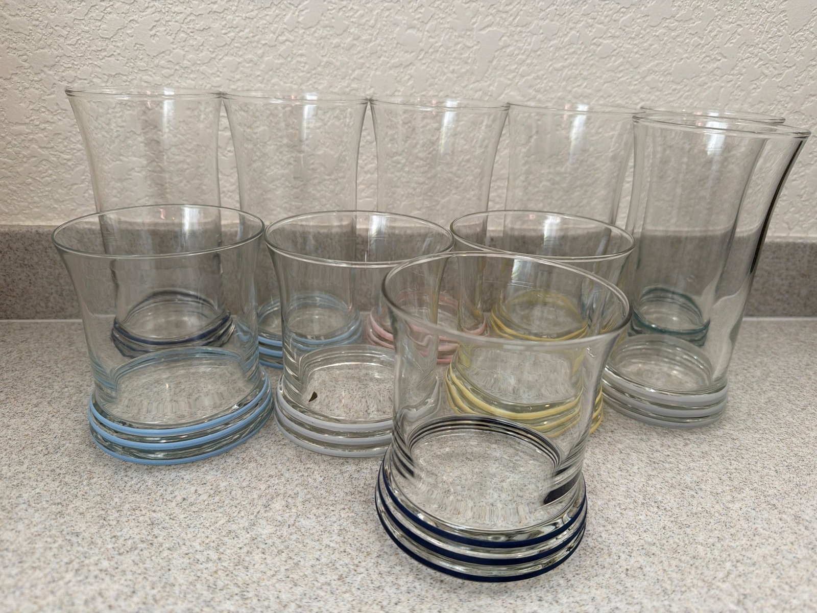 Colored Rim Glassware, Tumblers and Highball Glasses: Clear glass tumblers and highball glasses featuring colored rims in shades of blue, yellow, pink, and white. Pieces are made of glass and exhibit a uniform design with circular lines along the base. T