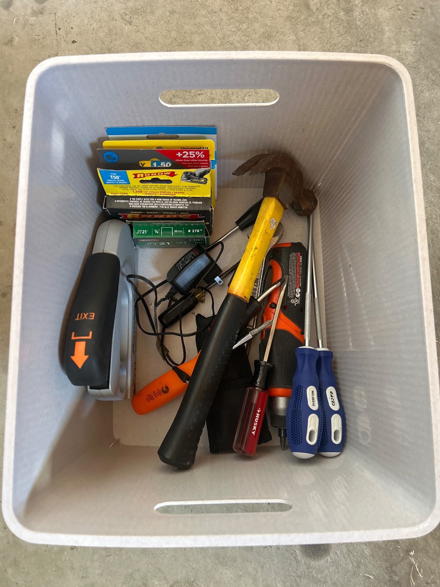 Plastic Storage Bin with Assorted Tools: A plastic storage bin containing assorted tools such as screwdrivers, a hammer, utility knife, electrical items, and a staple gun. Tools are gently used, suitable for various household tasks. Storage