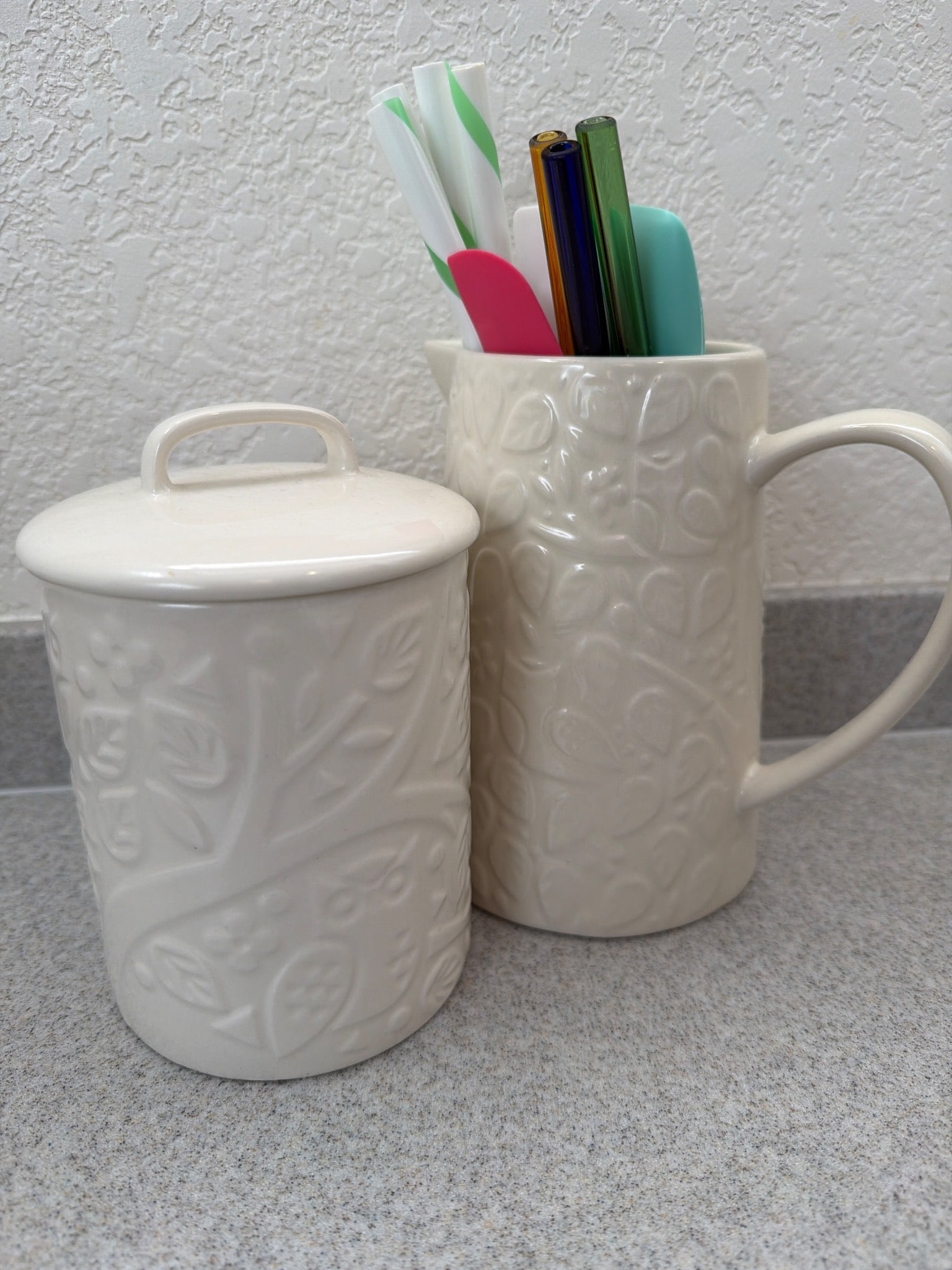 Mason Cash Embossed Ceramic Sugar Jar and Pitcher Set: Mason Cash ceramic set including an embossed sugar jar with lid and a pitcher, both featuring a delicate floral design. Made of durable ceramic material in a neutral finish. The base of each piece is