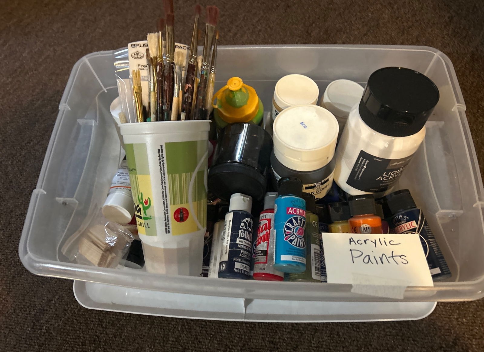 Acrylic Paints and Brushes in Plastic Storage Container: A plastic storage container filled with a variety of acrylic paints in assorted colors and sizes, along with an assortment of paintbrushes. Includes bottles of paint from various brands, yellow squeez