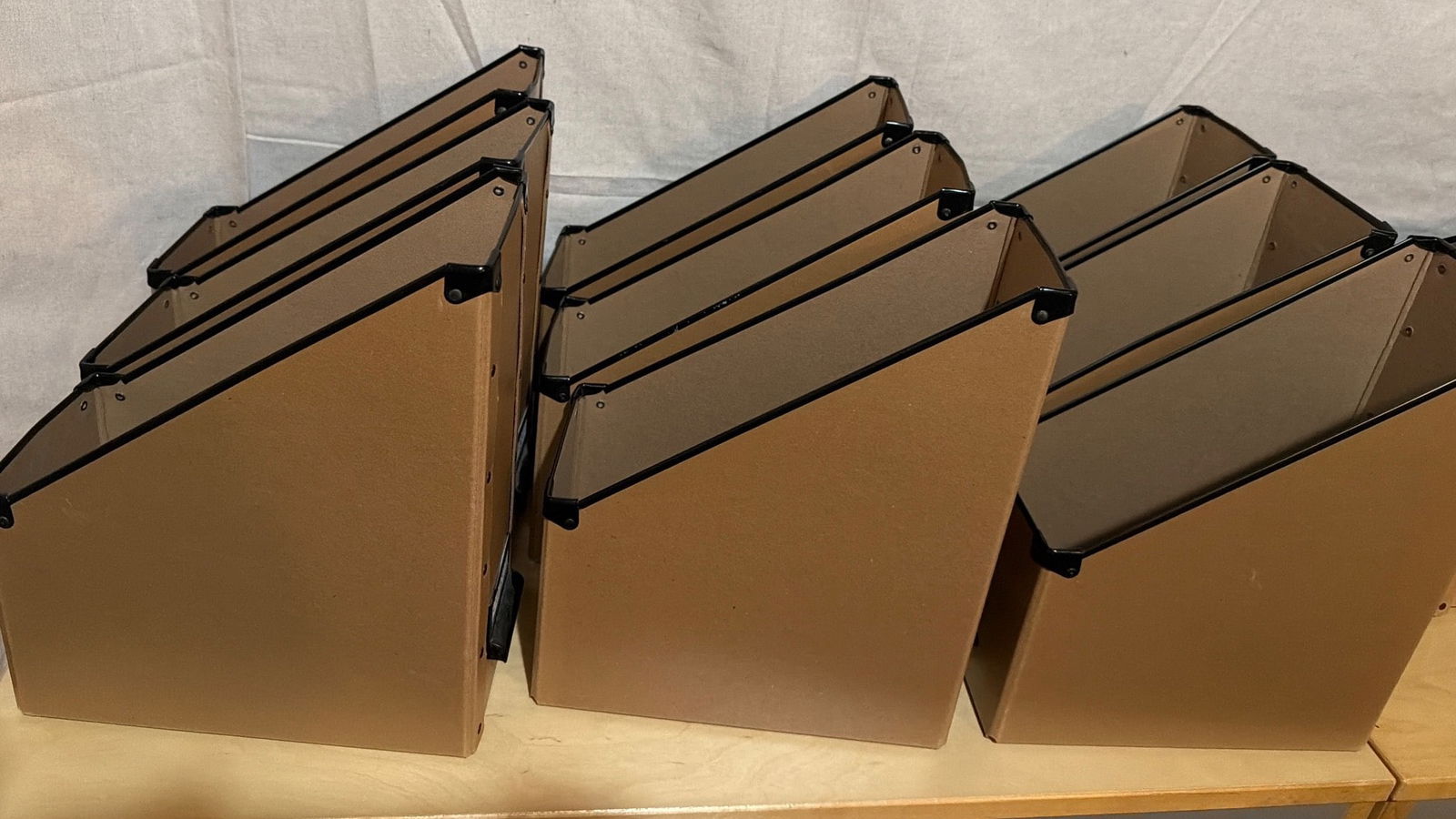 Six Brown Cardboard Magazine Holders With Black Reinforcements: Six cardboard magazine holders in brown with black plastic reinforcements on the corners. Constructed with a slanted opening for easy access to contents. Ideal for organizing documents, magazines, or