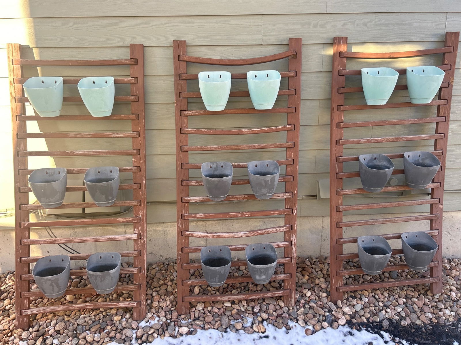 Three Wooden Plant Racks With Plastic Pots: Three wooden plant racks, each featuring multiple levels of slats and holding a combination of light green and black plastic pots. The racks have a weathered appearance, suggesting outdoor use. Each r