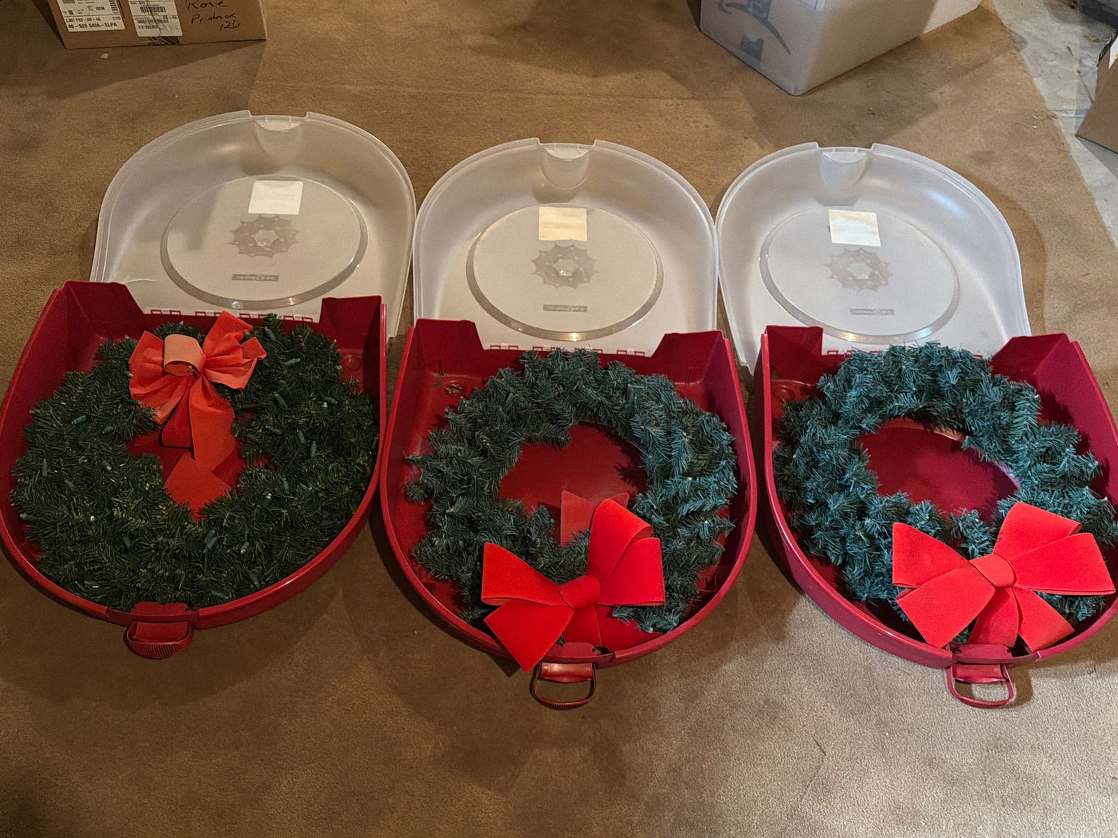 Three Decorative Holiday Wreaths with Storage Cases: Three artificial holiday wreaths, each adorned with red bows, and housed within red protective storage cases. The wreaths are constructed of greenery and feature durable designs. Ideal for seasonal de