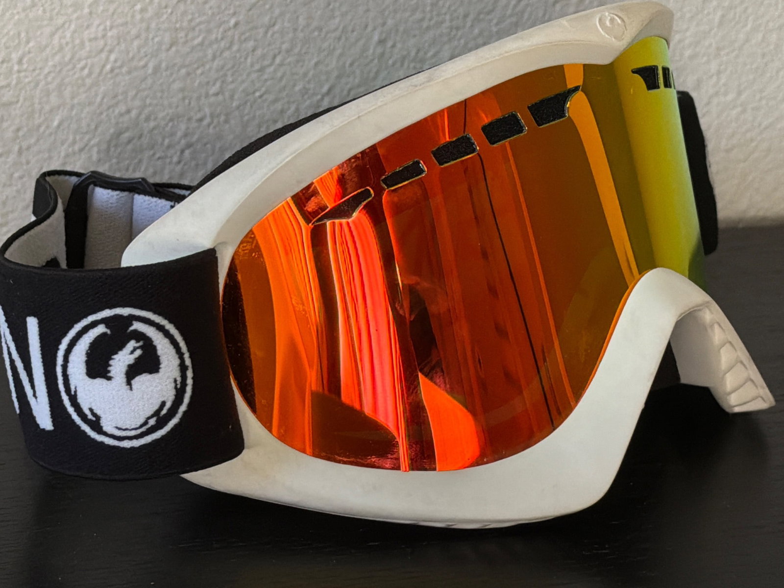 Dragon Snow Goggles With Orange Mirrored Lens: Dragon snow goggles featuring an orange mirrored lens and black adjustable strap with branding. The frame is white, and the goggles are equipped with foam padding for comfort. The strap includes a buc