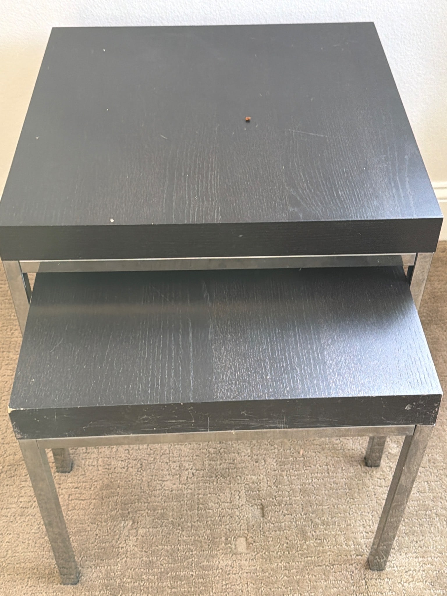 Two Modern Black Nesting Tables with Metal Base: Two black nesting tables featuring a modern design. The tables have square wooden tops with a black finish and are supported by sturdy metal legs. The tops show visible surface wear and a small hole i