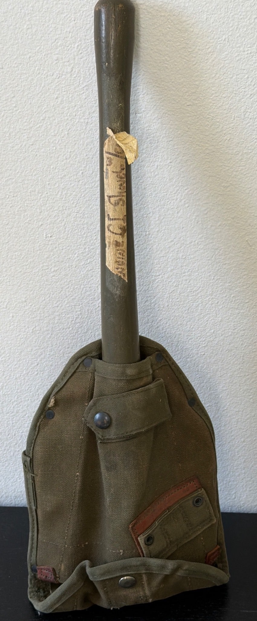 Vintage Soviet-Era Military Shovel with Canvas Cover: Vintage Soviet-era military shovel featuring a sturdy wooden handle and a detachable canvas cover. The tool exhibits signs of wear, consistent with its age and usage, including remnants of a partially
