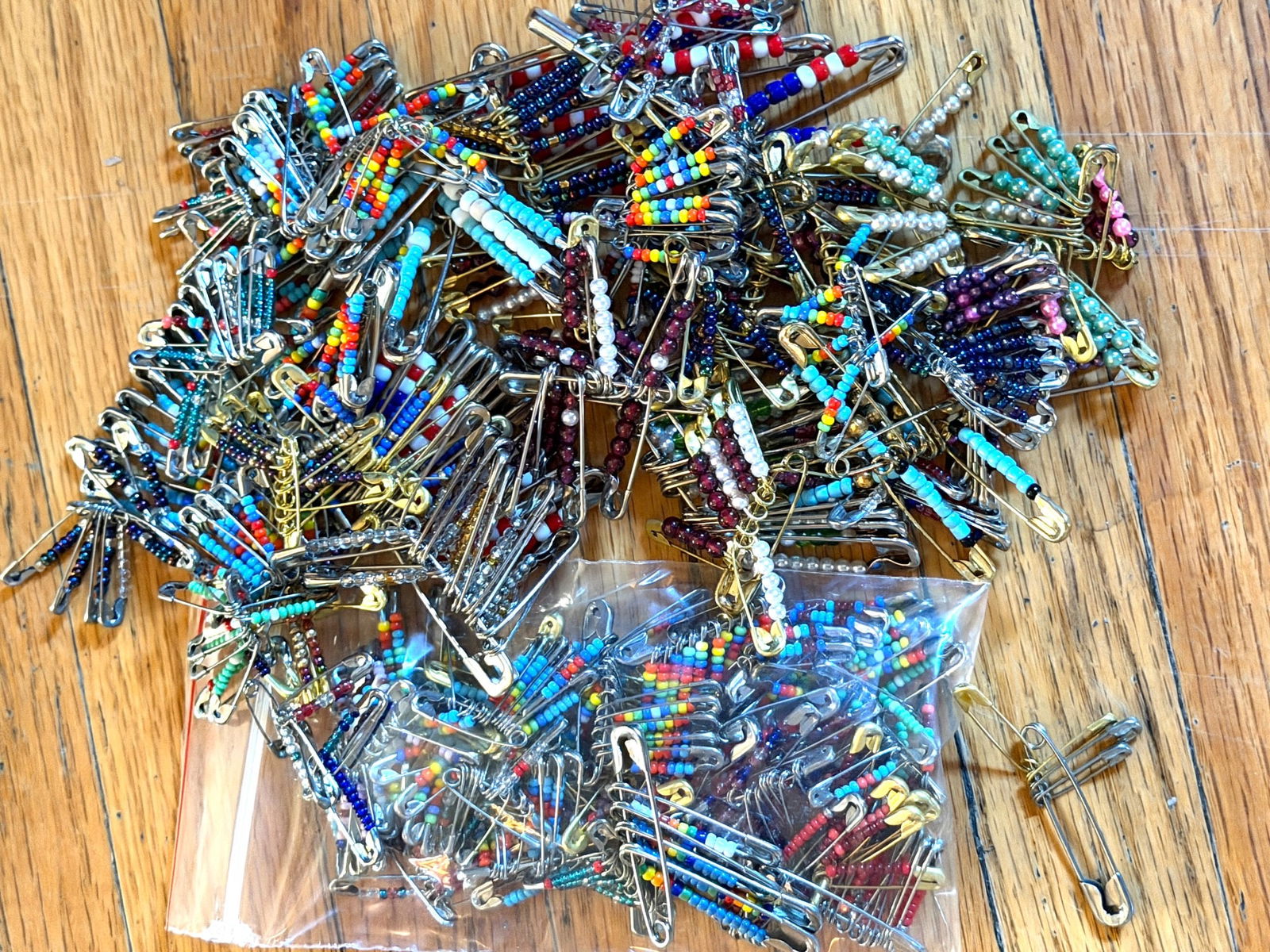 Beaded Safety Pin Crafts in Various Designs and Colors: Safety pin crafts featuring colorful glass beads in various designs and patterns. The lot includes multiple beaded creations, some with intricate patterns such as flags, geometric shapes, and abstract