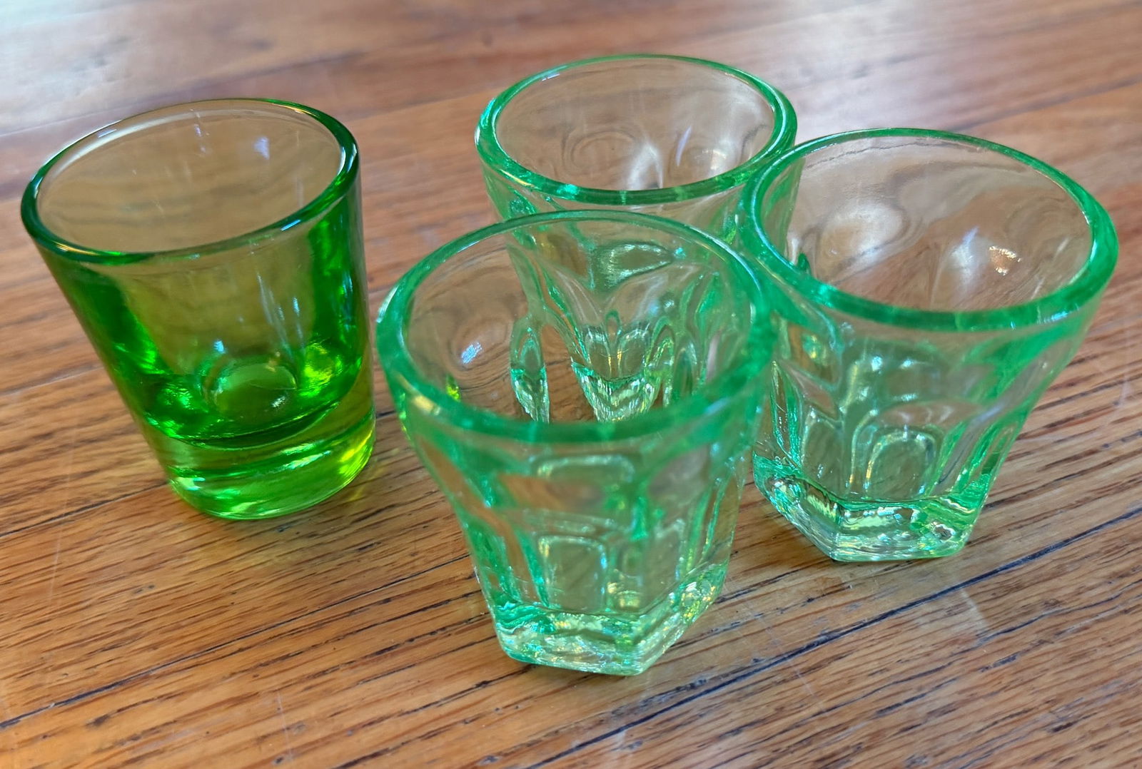 Four Green Glass Shot Glasses, Hexagonal Bases: Four green glass shot glasses featuring hexagonal bases and smooth finishes. Glasses display a uniform green tint and clear detailing. Approximate height of each glass is 2 inches, with a base diamete