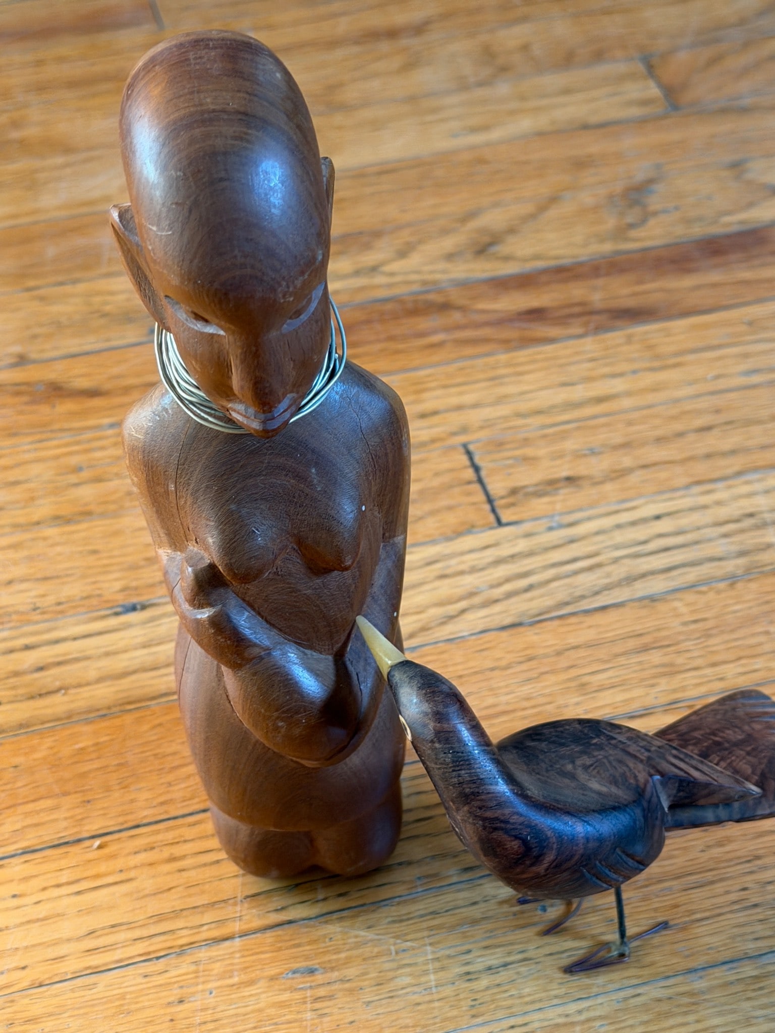 Wood Carved Sculpture of Woman and Bird with Metal Accents: Hand-carved wooden sculpture depicting a standing woman interacting with a bird. The figure has detailed carving on the face and body, and the bird is connected to the woman in a feeding pose. The scu