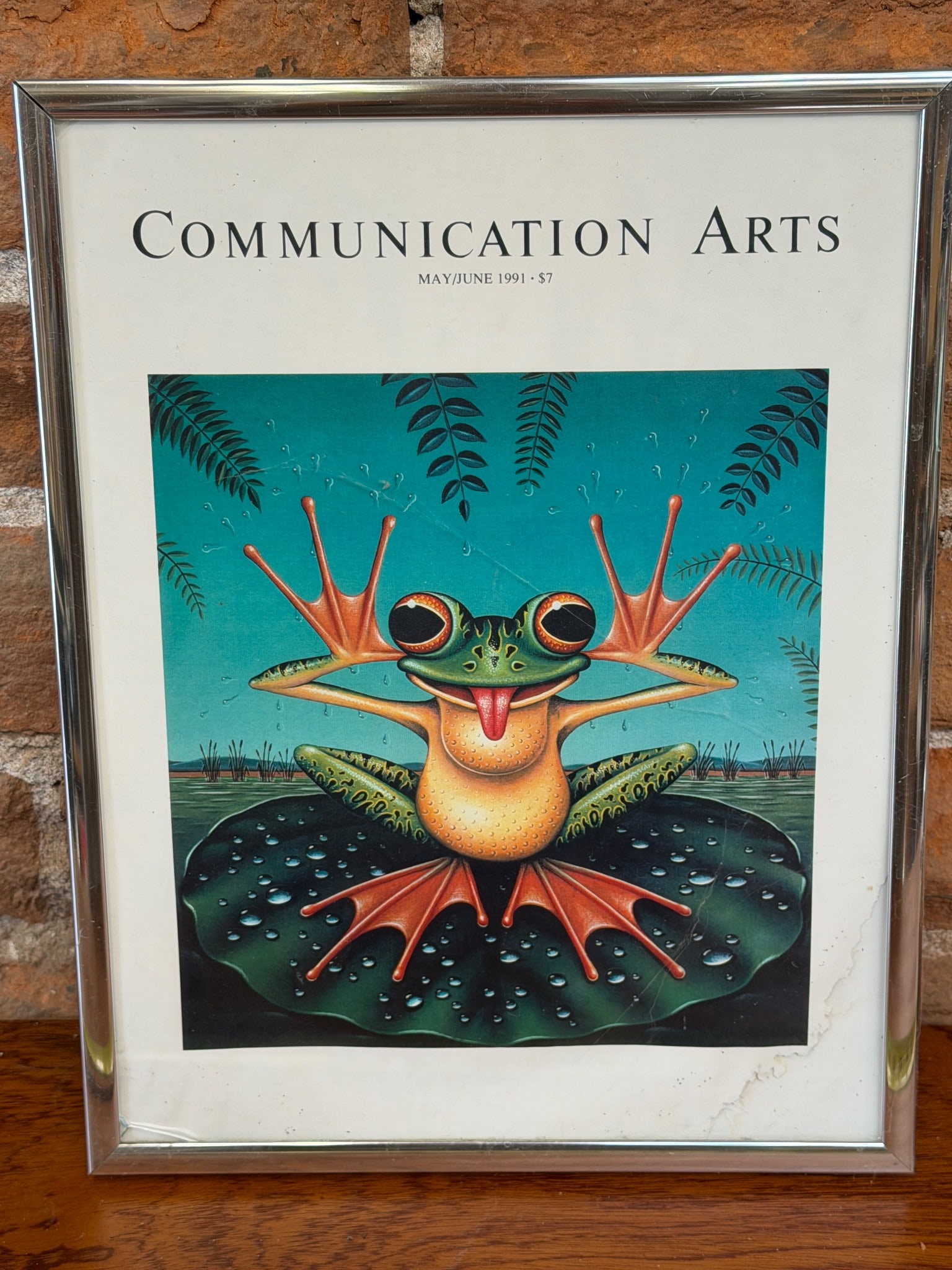 Communication Arts Cover Artwork in Metal Frame, November 1987: Framed cover artwork from the November 1987 issue of Communication Arts Magazine featuring a colorful illustration of a frog. The artwork is encased in a metal frame with visible wear, including a cor