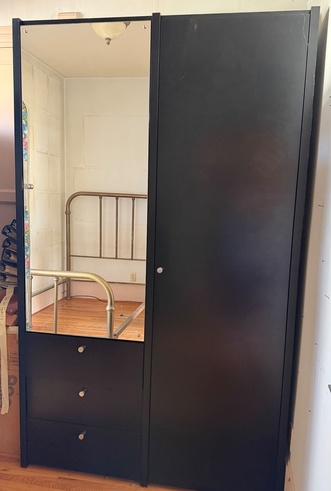 Black Armoire With Mirrored Door and Drawers: Black armoire featuring a mirrored door, hanging rod, interior shelf, and two bottom drawers. The unit is made of wood or composite material and includes metal hardware. Dimensions are approximately 8