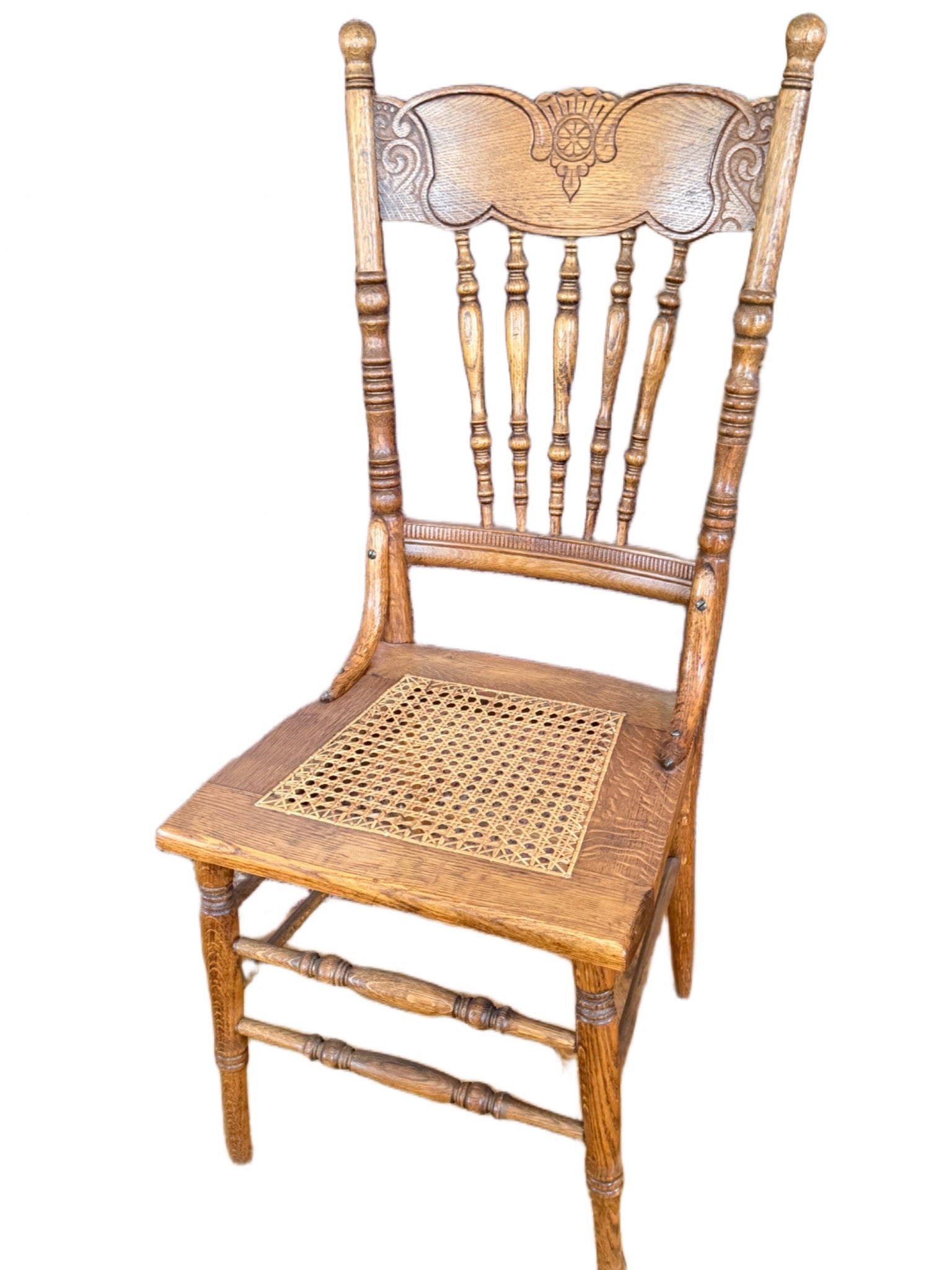 Ornate Oak Chair with Caned Seat and Decorative Backrest: Ornate oak chair featuring an intricately carved decorative backrest, turned spindles, and a woven cane seat. The backrest displays detailed carvings, while the legs and stretchers exhibit turned wood