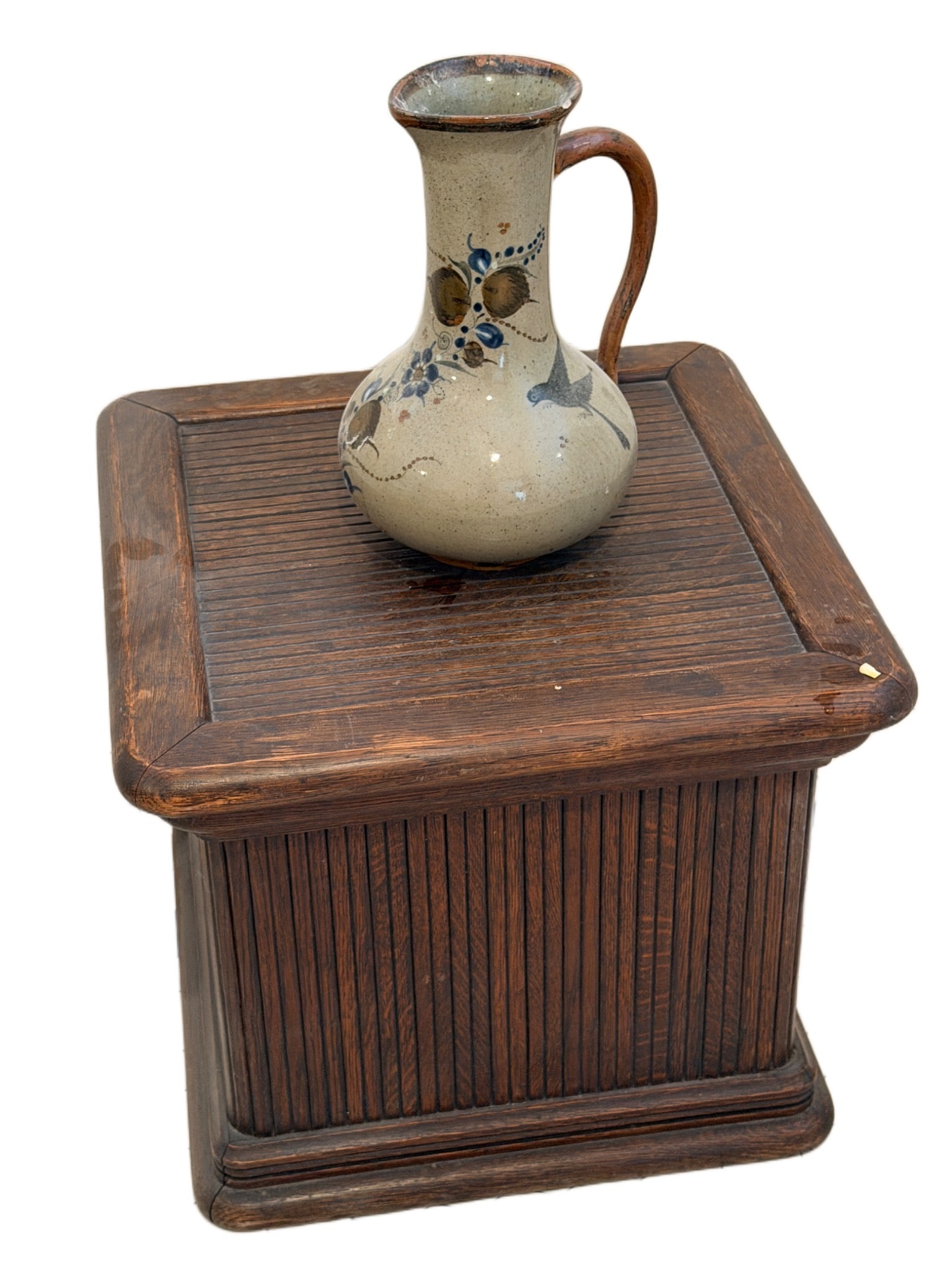 Handcrafted Ceramic Jug Signed by Artist with Wooden Table Base: Handcrafted ceramic jug featuring a painted floral and bird motif, signed by the artist on the underside. The jug shows signs of use, including surface wear and a chip on the rim. It measures