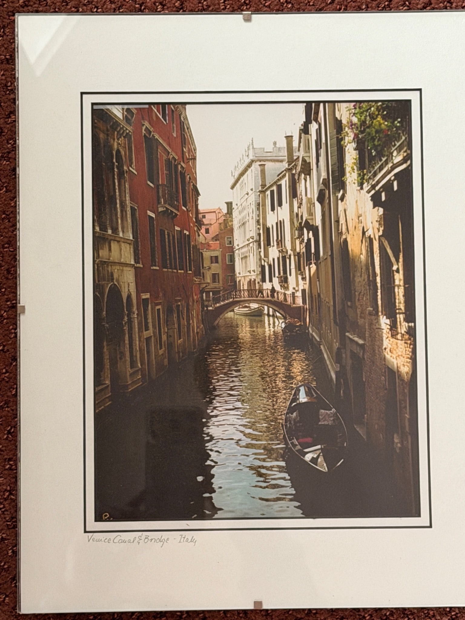 Framed Photograph of Venice Canal and Bridge, Italy: Framed photograph titled 'Venice Canal & Bridge - Italy,' featuring a tranquil canal scene with reflection and historical architecture. The photograph is mounted on a white mat and secured on a backin