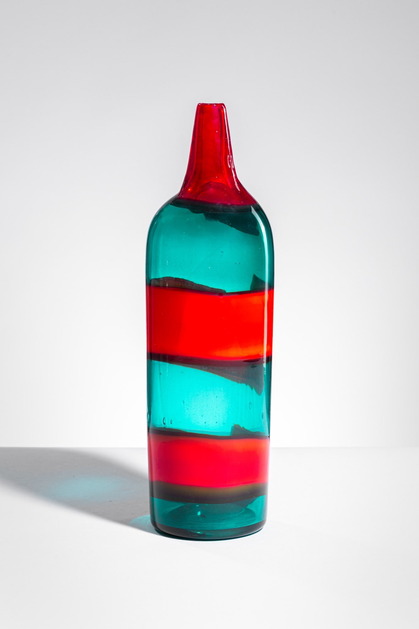 Fulvio Bianconi. Bottiglia della serie a Fasce Orizzontali mod. 4315: Bottiglia della serie a Fasce Orizzontali mod. 4315 Vetro soffiato trasparente verde, decorato sul corpo da due fasce orizzontali in rosso opaco e sul collo da una fascia in rosso trasparente. Venini,