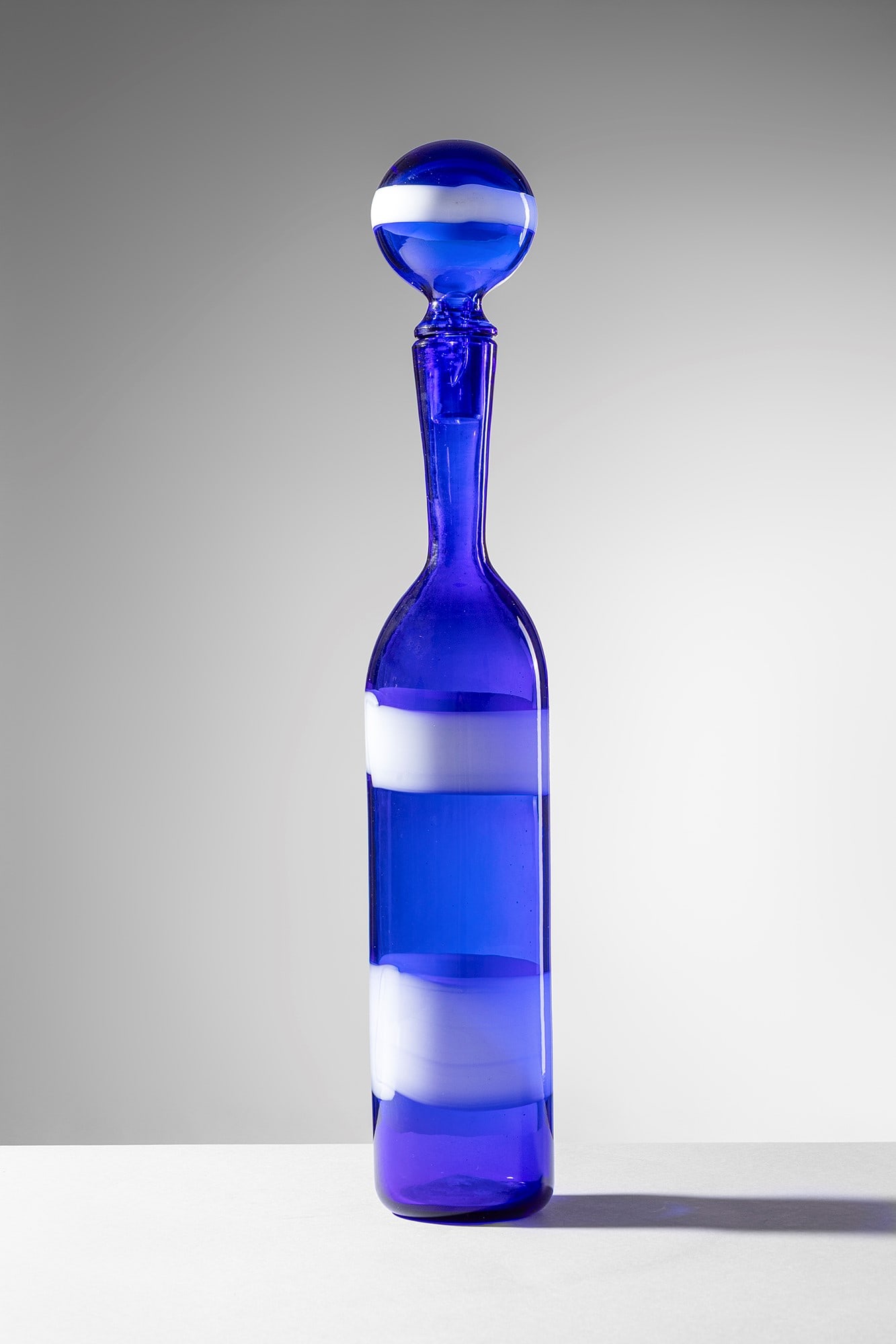 Fulvio Bianconi. Bottiglia a fasce orizzontali mod. 4582: Bottiglia a fasce orizzontali mod. 4582 Vetro soffiato blu decorato da fasce orizzontali lattimo. Venini, 1953-56. Firma all'acido circolare Venini Italia. h cm 47, diam. cm 9 Bibliografia: Marino Bar