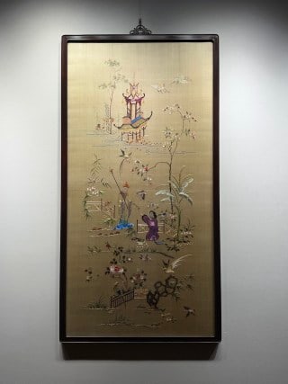 A picture of the ceremony of the sage of Guangdong embroidery in the Republic of China: Vertical vertical axis, beige silk ground, weaving and painting courtyard scenery, pavilions, plantain flowers and trees, people holding things in the courtyard, surrounded by cranes and birds, the