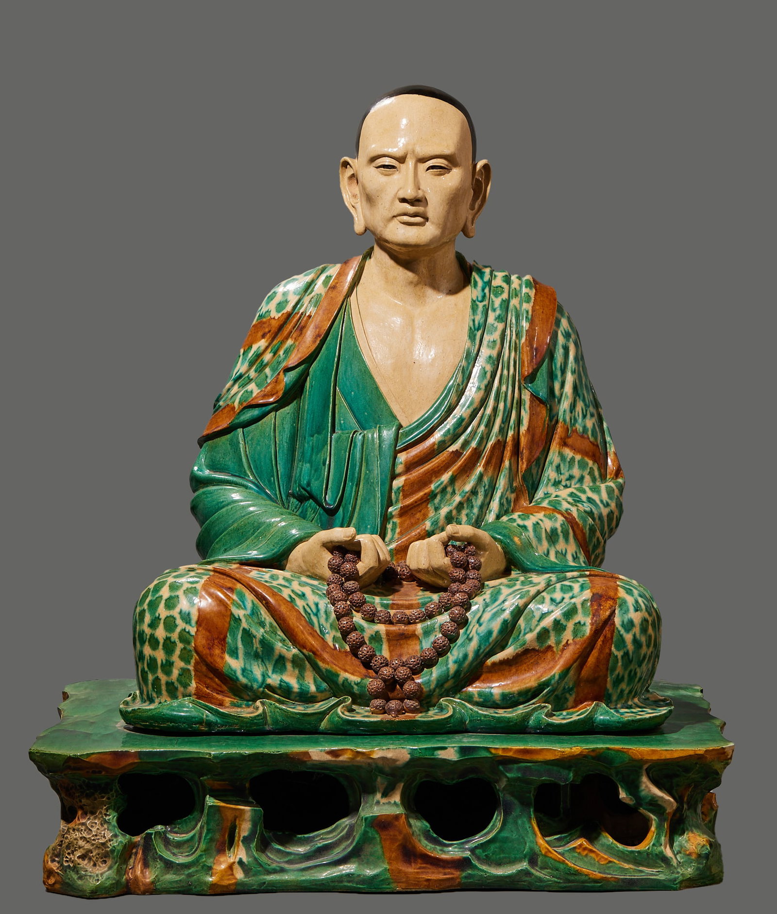 Sitting statue of arhat before Ming Dynasty: Lohan is in a posture of squatting, with a realistic face, bald head and wide ears. Hold a string of bodhi beads in front of your abdomen with both hands. Wearing a multi-layer monk's robe, the glaze