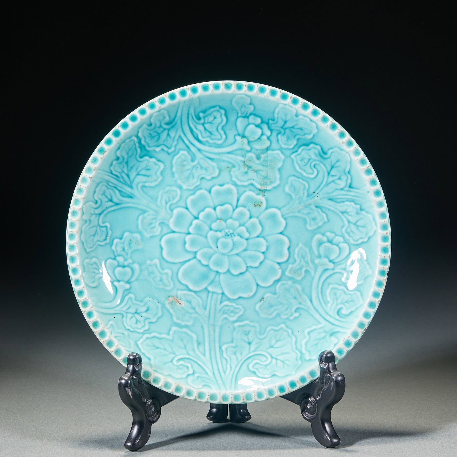 Blue glazed flowered plate: Round porcelain plate with turquoise glaze. The center of the disc is printed with a large peony, which extends outward with branches and leaves. The dish is decorated with a circle of beads. The over