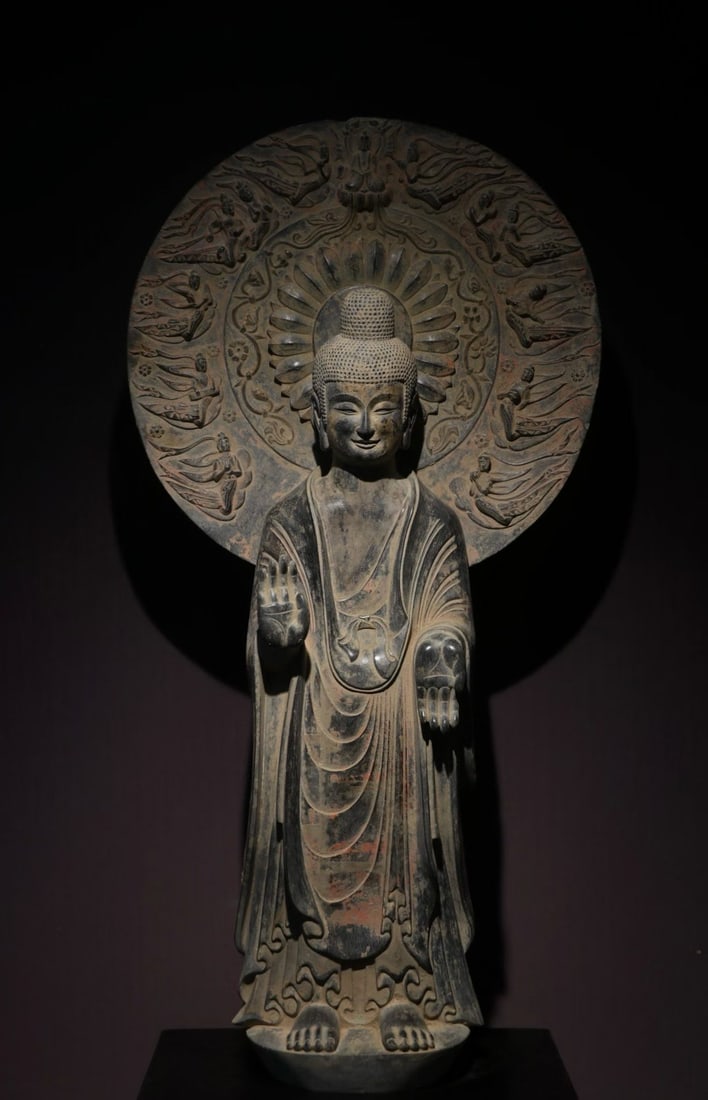 Gold-plated flying backlit Buddha statue: A standing statue of Buddha made of bluestone, with a plump bun and a smiling face. Wearing a draped robe, the robes are layered smoothly, and there are traces of red color left in the robes. The