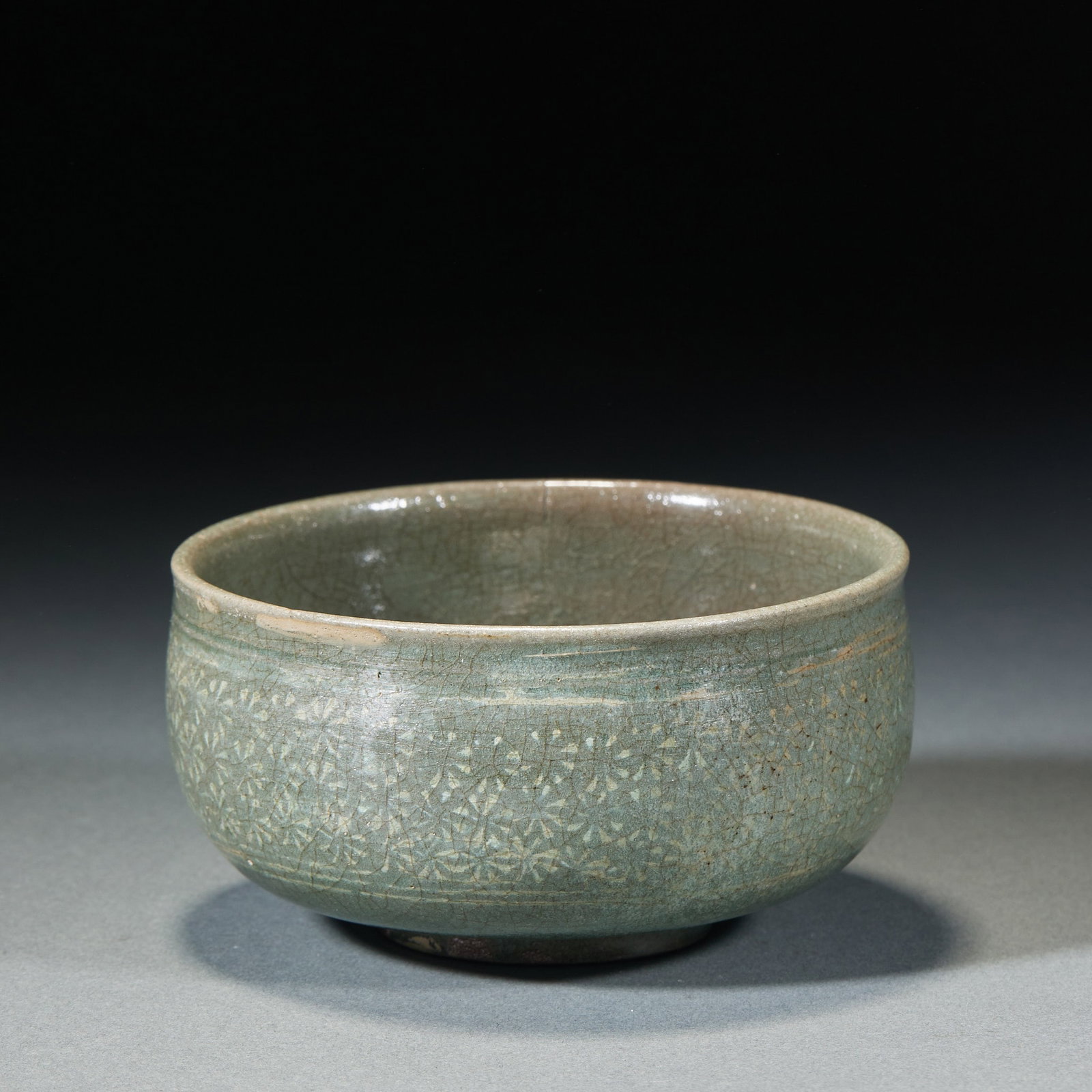 Korean celadon flower pattern bowl: Open, arc belly, circle foot. The whole body is painted with green and gray glaze, and the glaze is spread all over. The outer wall of the bowl is stamped with a circle of continuous floral patterns,