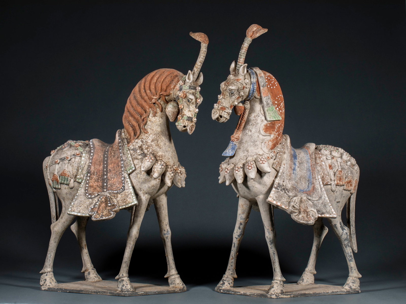 Pair of Pre-Ming Pottery War Horses: Of earthenware retaining red, green, white and blue painted decoration. The two horses stand facing each other, both with saddlery and pendant ornaments, long tassels on their heads; details of the