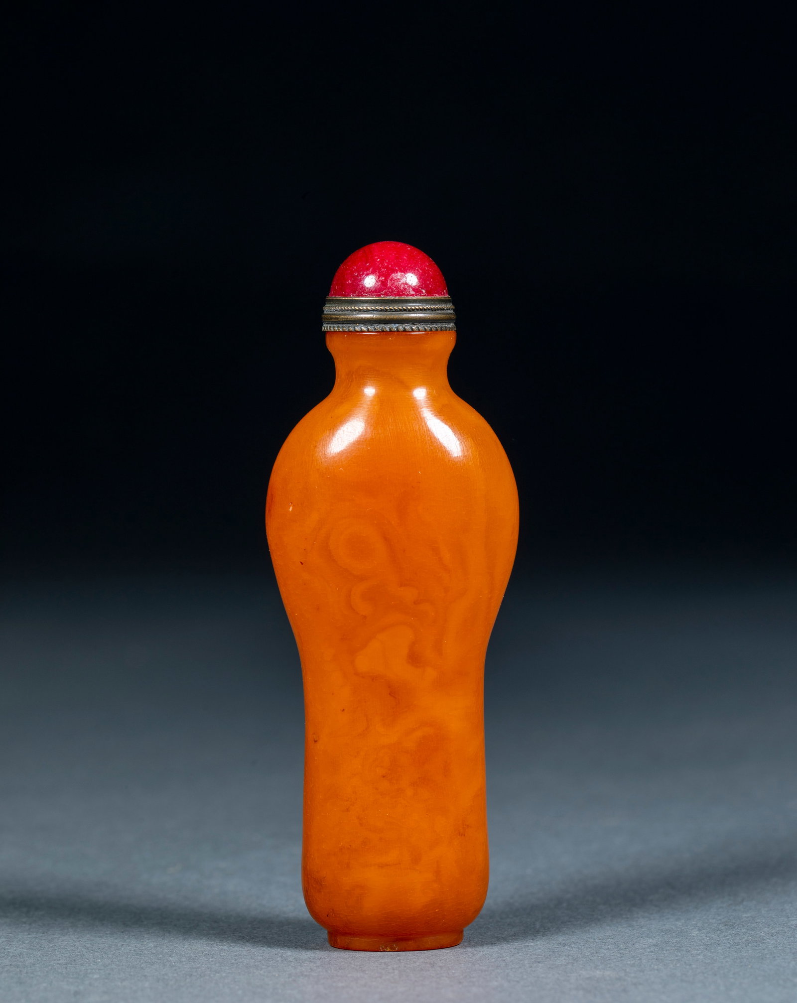 Amber Snuff Bottle: Of orange-yellow amber with natural flowing grain, bottle-shaped, with a threaded metal mouth and a red domed stopper. H 9.3 cm, L 3.3 cm, W 2.2 cm, 37.5 g.