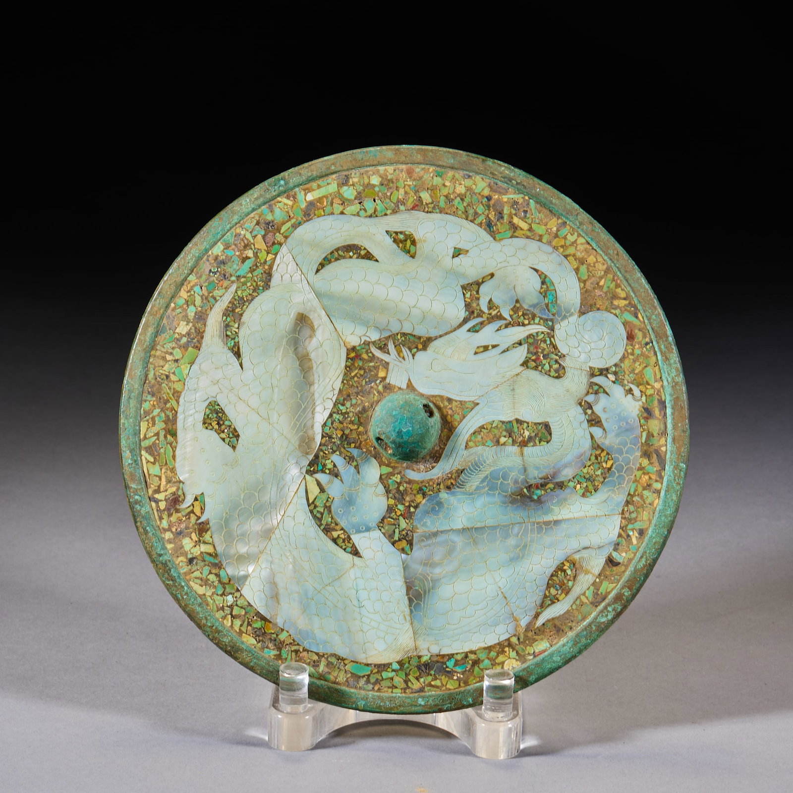 Mother-of-Pearl Inlaid Bronze Mirror with Coiled Dragons: A circular bronze mirror; the back is inlaid in mother-of-pearl with twin dragons on a ground densely set with turquoise chips; the knob is of verdigris copper. Diameter 20.5 cm.