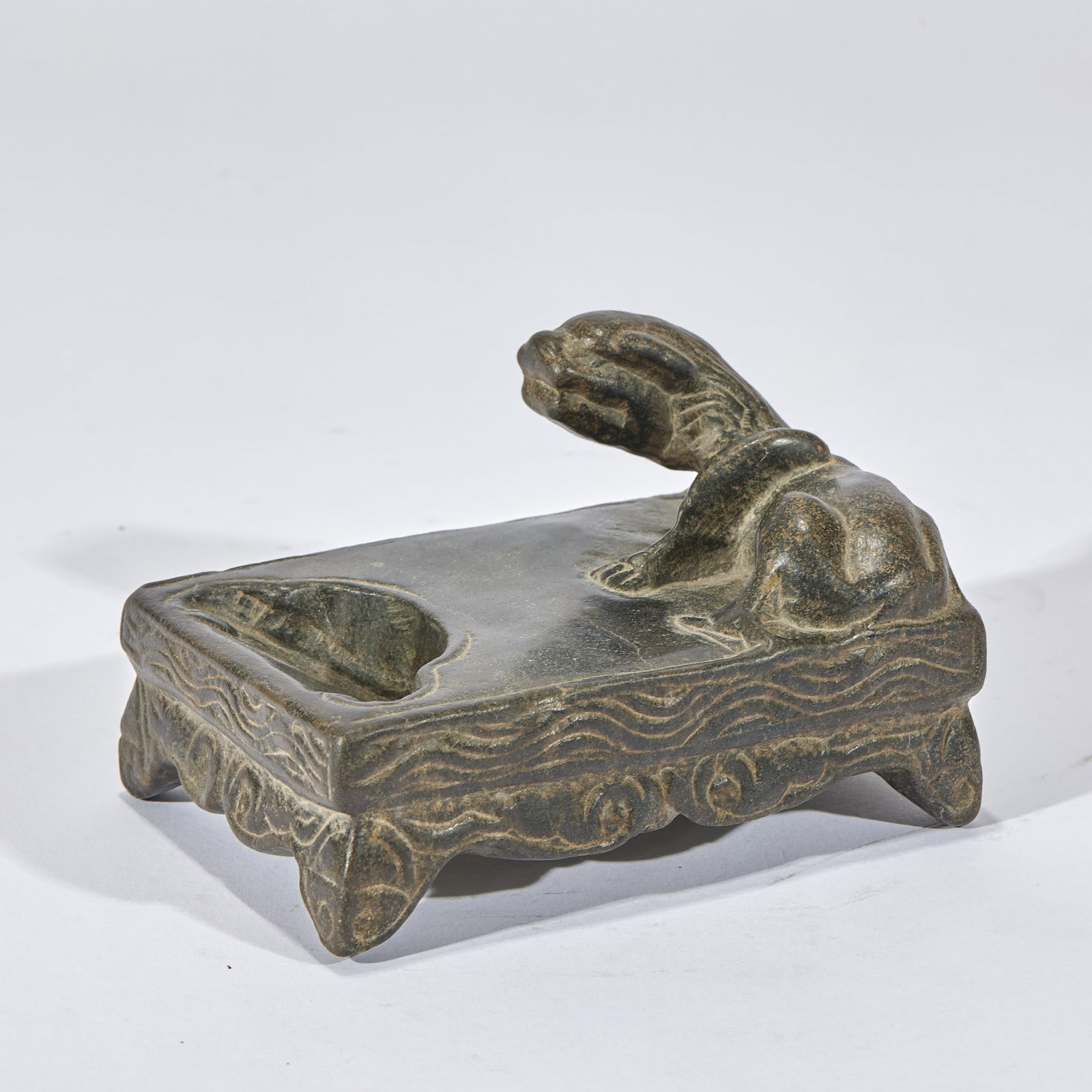 Stone Auspicious-Beast Inkstone: A rectangular blackish-green stone inkstone; one end opens an ink pool and a reclining beast is carved in high relief at the upper right. The sides are carved with wave patterns and it stands on four