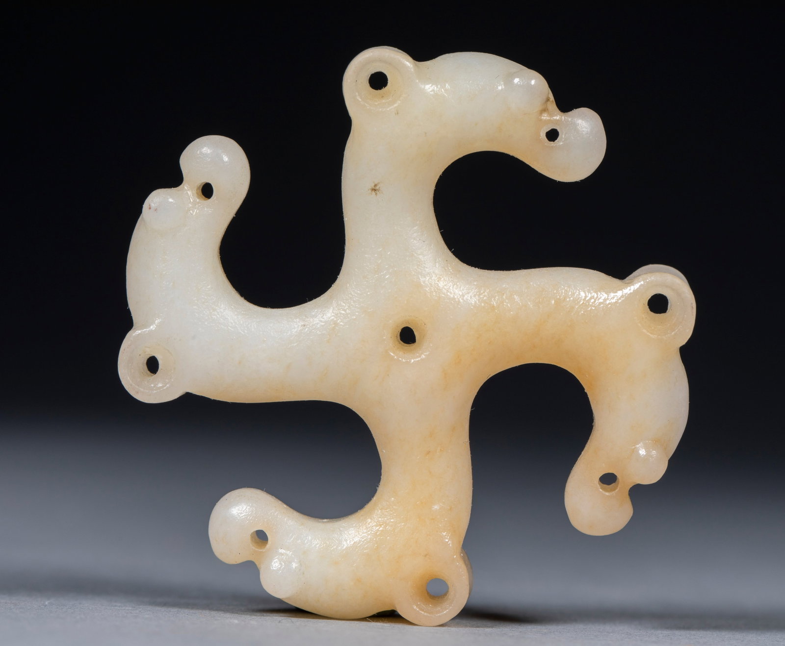 Jade Object of Four Beast Heads: Of white jade with light-yellow calcification, in a swastika-like form assembled from dragons linked head to tail, covered with perforations for stringing into group pendants. Unique in form ?? a