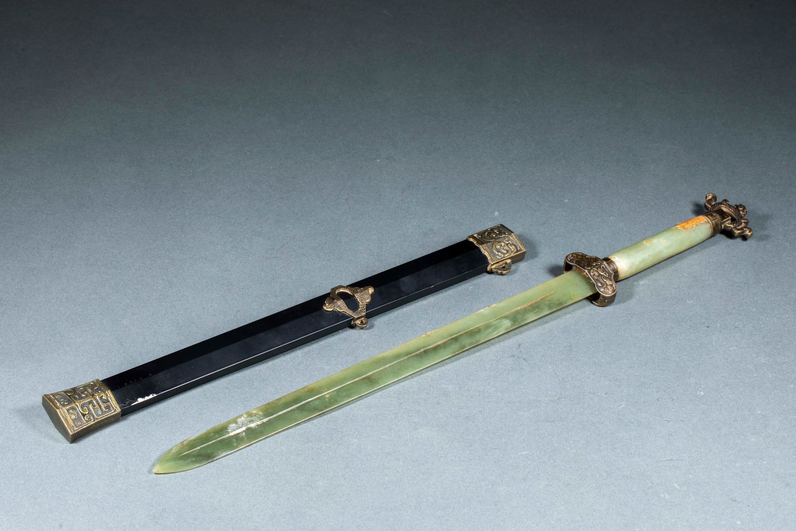 Hetian Jade Sword: The blade is carved from greenish jade with old-copper fittings of guard, pommel and scabbard-mounts ornamented with kui-dragons; the scabbard is black-lacquered. The jade is warm and the form