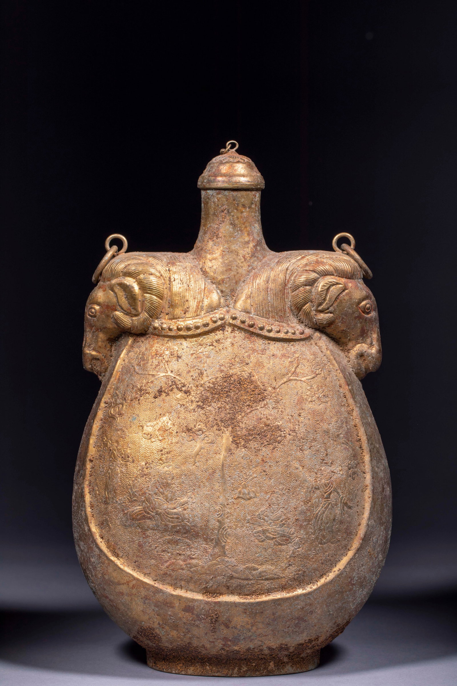Ancient Chinese Sheep-Head Leather Flask: With two horse-heads holding rings on the sides; the body is densely engraved with fish-roe ground and panels carved with a figure-story scene beneath a tree, with an original cover. The form derives