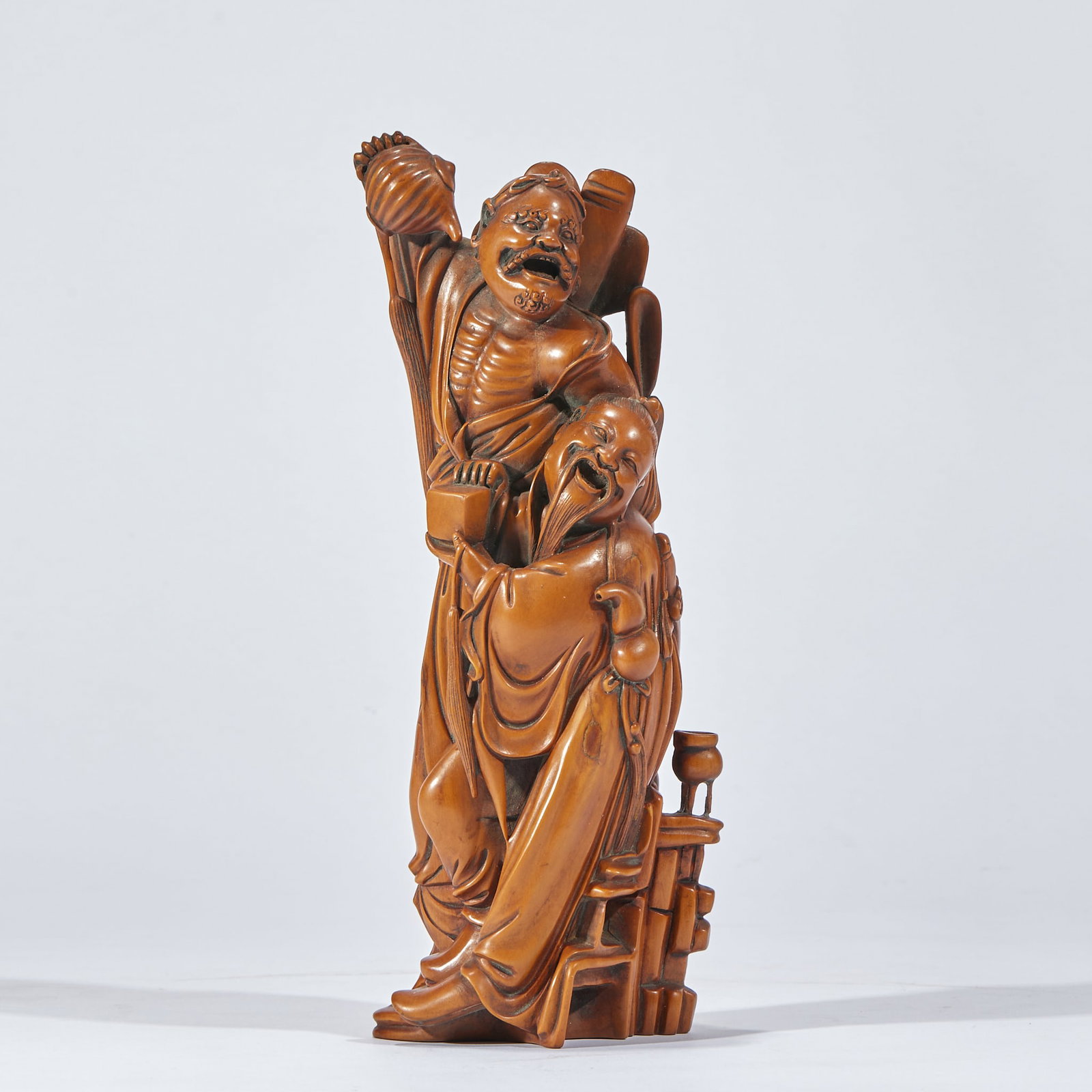 Boxwood Bodhidharma Figure: Carved from a single block of boxwood, two immortals leaning together ?? one raising an implement, one holding a treasure box, with a gourd and a musical instrument beside; robes flowing and