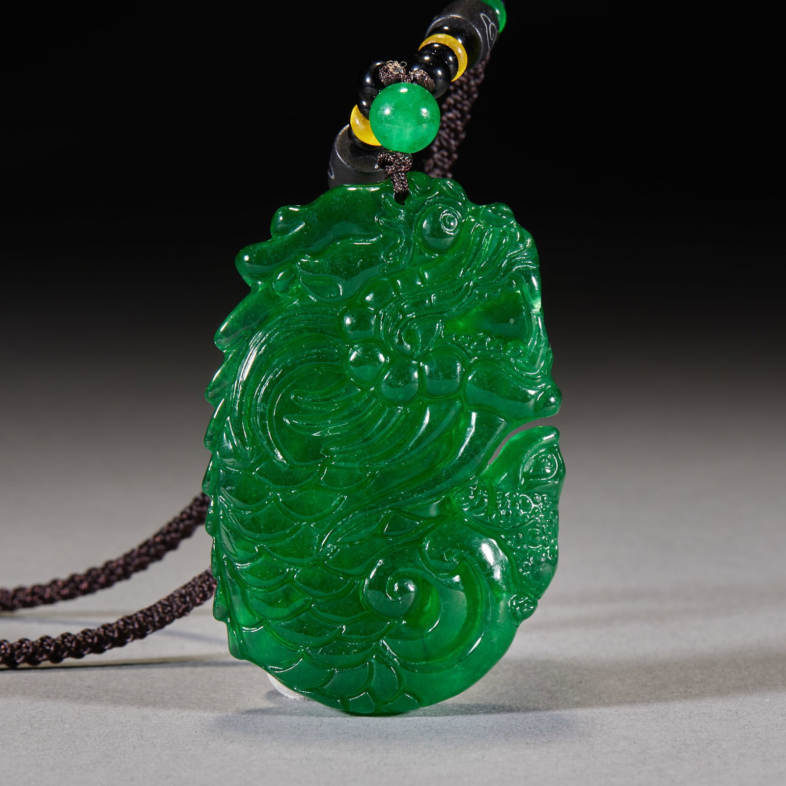 Jadeite Dragon Plaque: High-colour full-green jadeite carved with a fish-transforming-dragon motif: the fish body stretches and the dragon head rises, richly layered carving. Strung on a multi-treasure woven cord; the