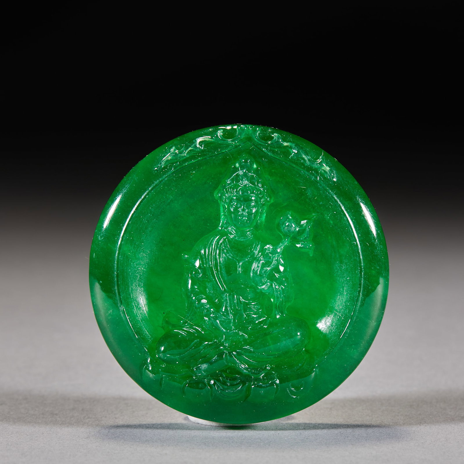 Jadeite Guanyin Plaque: A round jadeite plaque of dense full green; the centre is carved in relief with a lotus-holding Guanyin seated in lotus position with a serene face; the rim is carved with scrollwork. The stone is