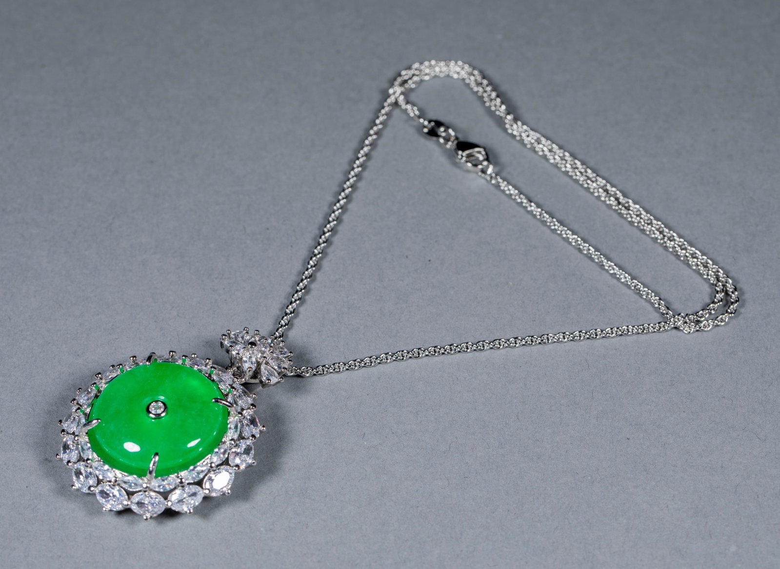 Jewelry Necklace: An ice-green full-green jadeite 'peace buckle' ringed by a silver frame set all around with diamonds, a central stone, with a double chain. The peace buckle form is complete and the setting sumptuous 