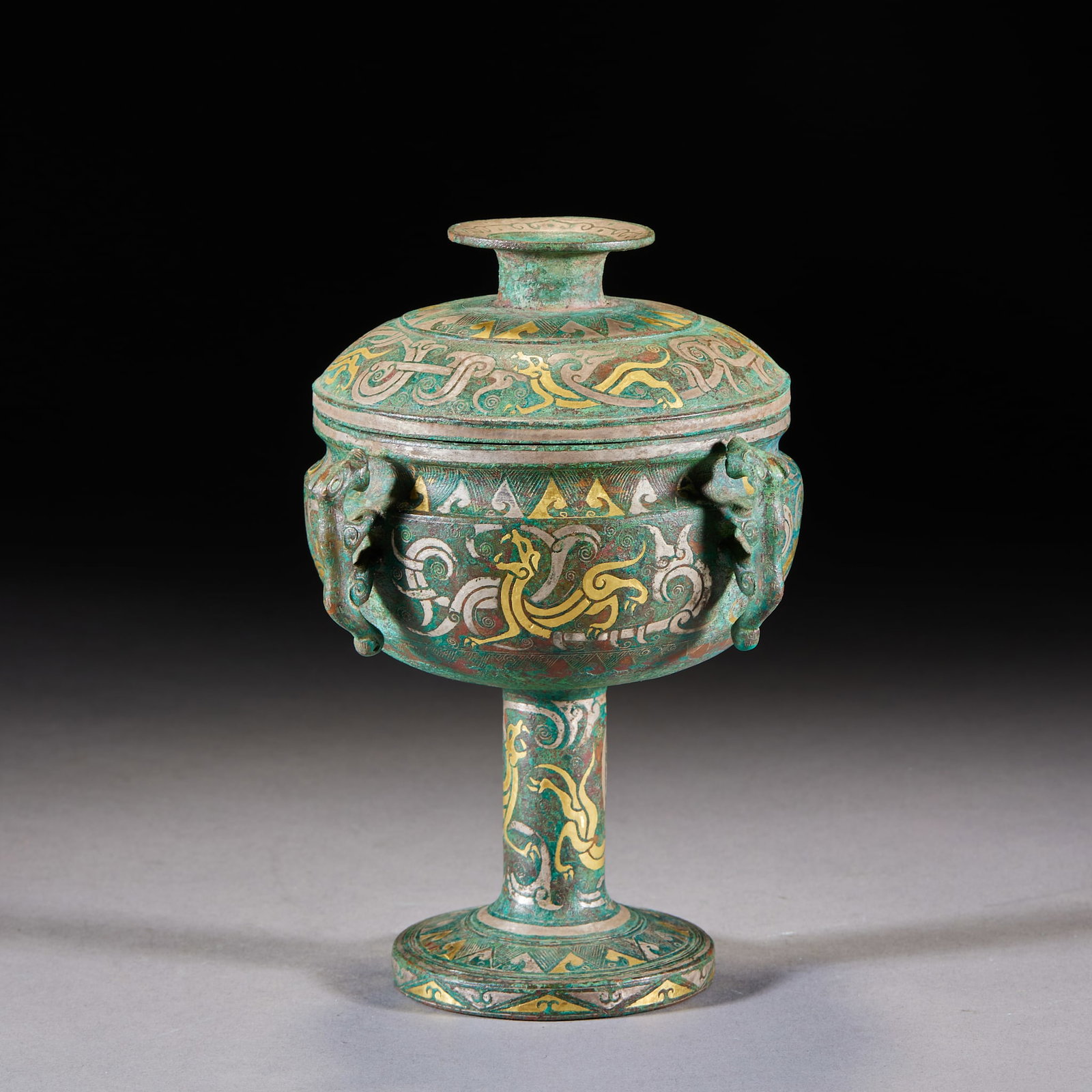 Inlaid-Gold-and-Silver Four-Beast-Ear Chi-Tiger High-Footed Covered Box: A bronze covered dou with tall stem and lid; body, lid and stem are inlaid throughout with gold and silver in dragon and cloud-scroll patterns, with beast-head ears at the sides and rich green patina