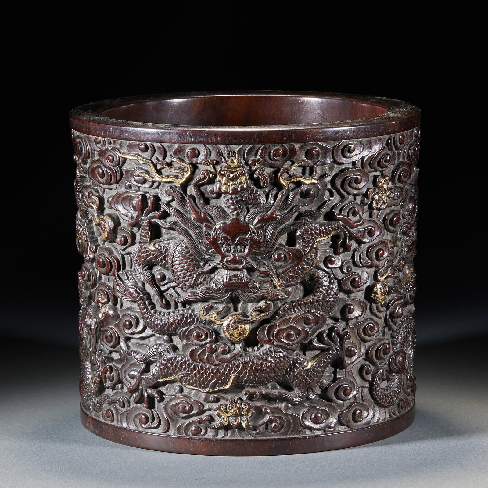 Zitan Carved Cloud-Dragon Brush Pot: A straight zitan brush pot with the exterior carved in relief with cloud-dragons coiling through a sea of clouds, teeth bared and claws out, imposing. The grain is fine with a warm patina and assured