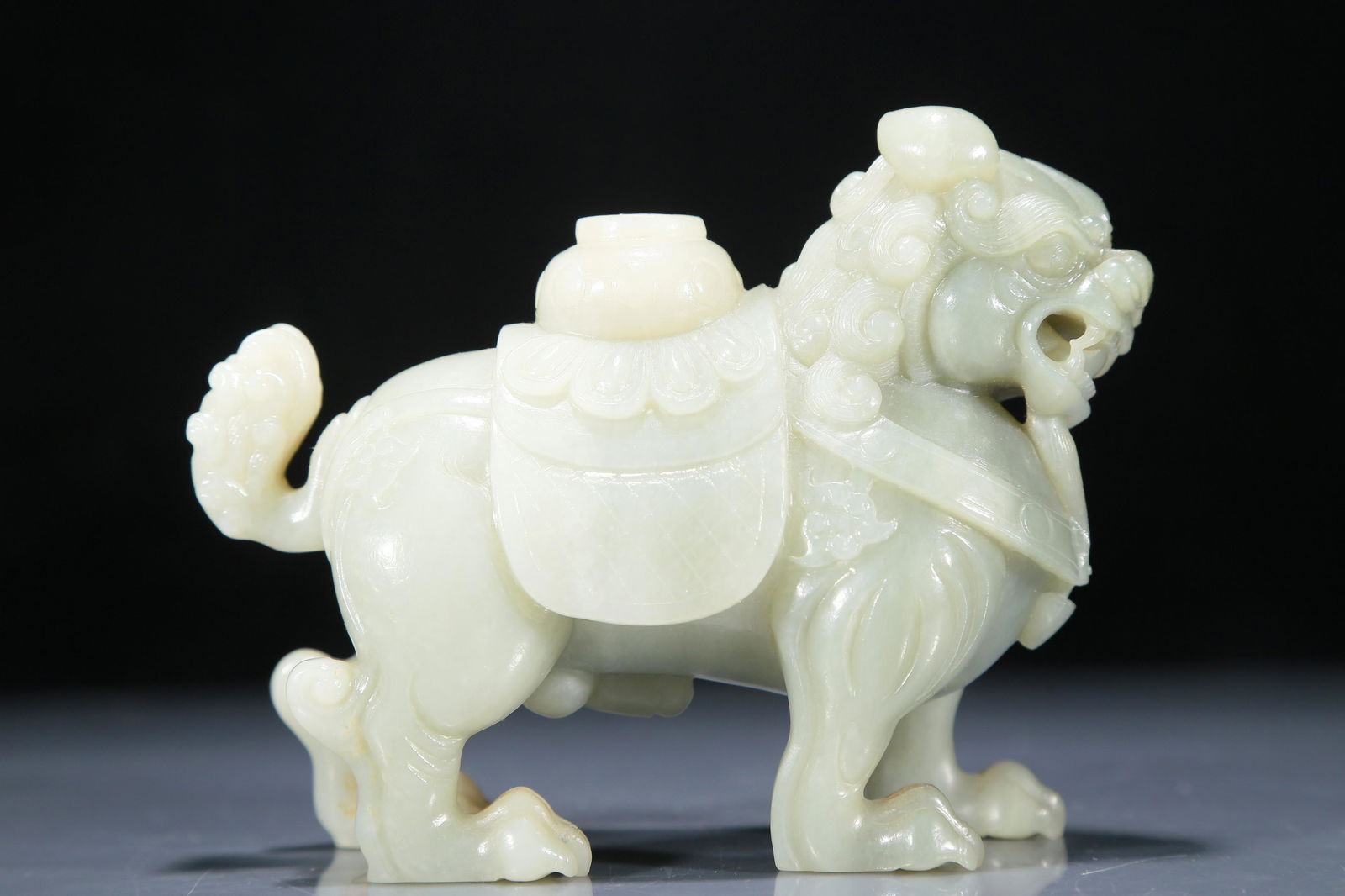 An Exquisite White Jade Lion: An Exquisite White Jade Lion. Standing four-square, the jade body caparisoned with a saddle draped with a cloth decorated with patterns, with a miniature jar rested atop on its back, mouth slightly op