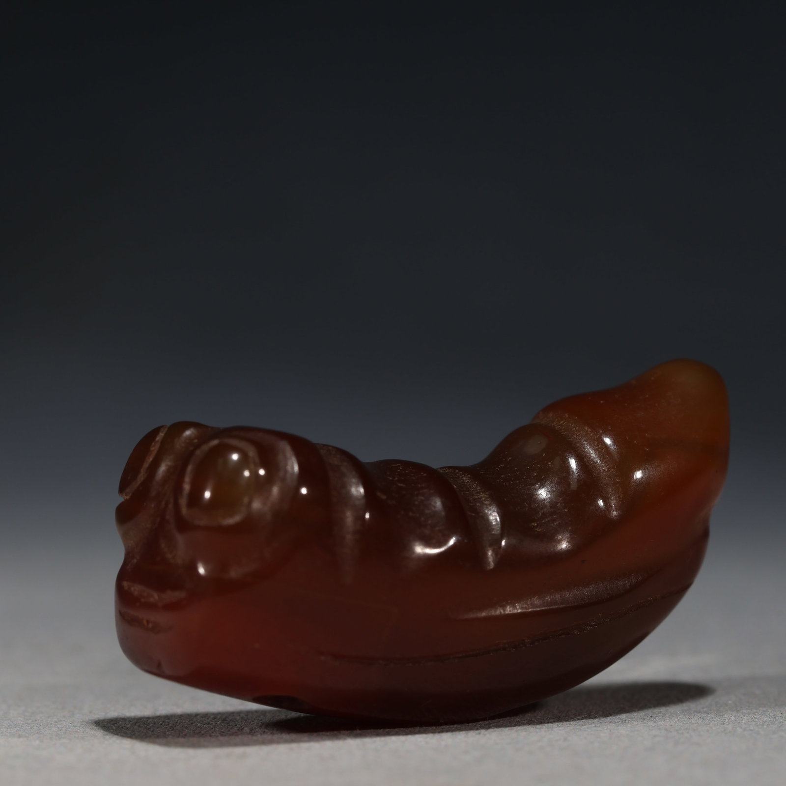 RED AGATE CARVED CHILONG PENDANT: A finely carved red agate pendant depicting a coiled mythical beast(chilong)with a rounded head,bulging eyes,and curved body.The stone is of a deep reddish-brown tone with a polished,glossy surface.&#