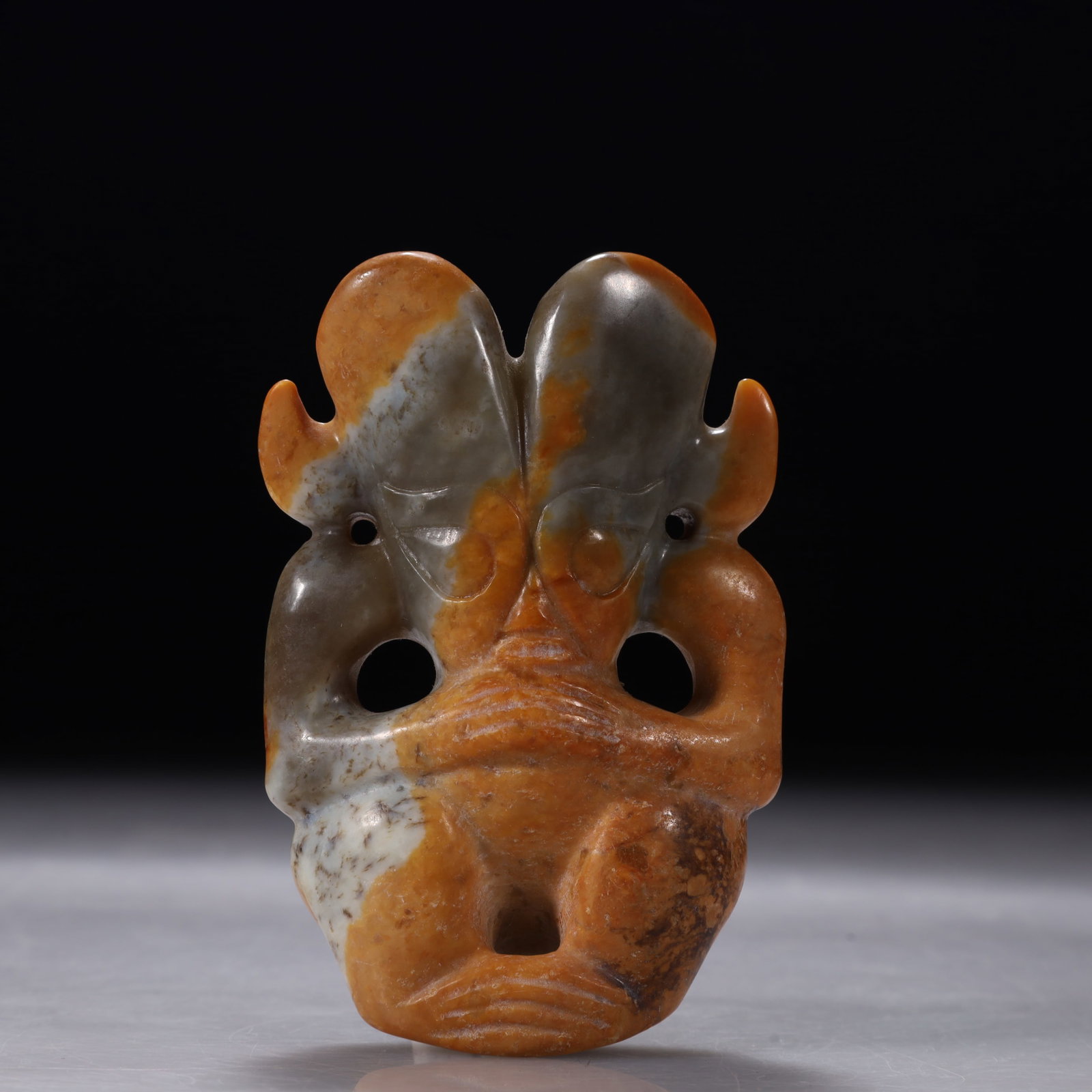 Hongshan Culture Jade Beast Face Pendant: Flat carving of a beast face with large round eyes, a wide snout, and two outward-curling horns on the head. Perforations are set at the bottom and jaw for suspension. The carving is archaic and abstr
