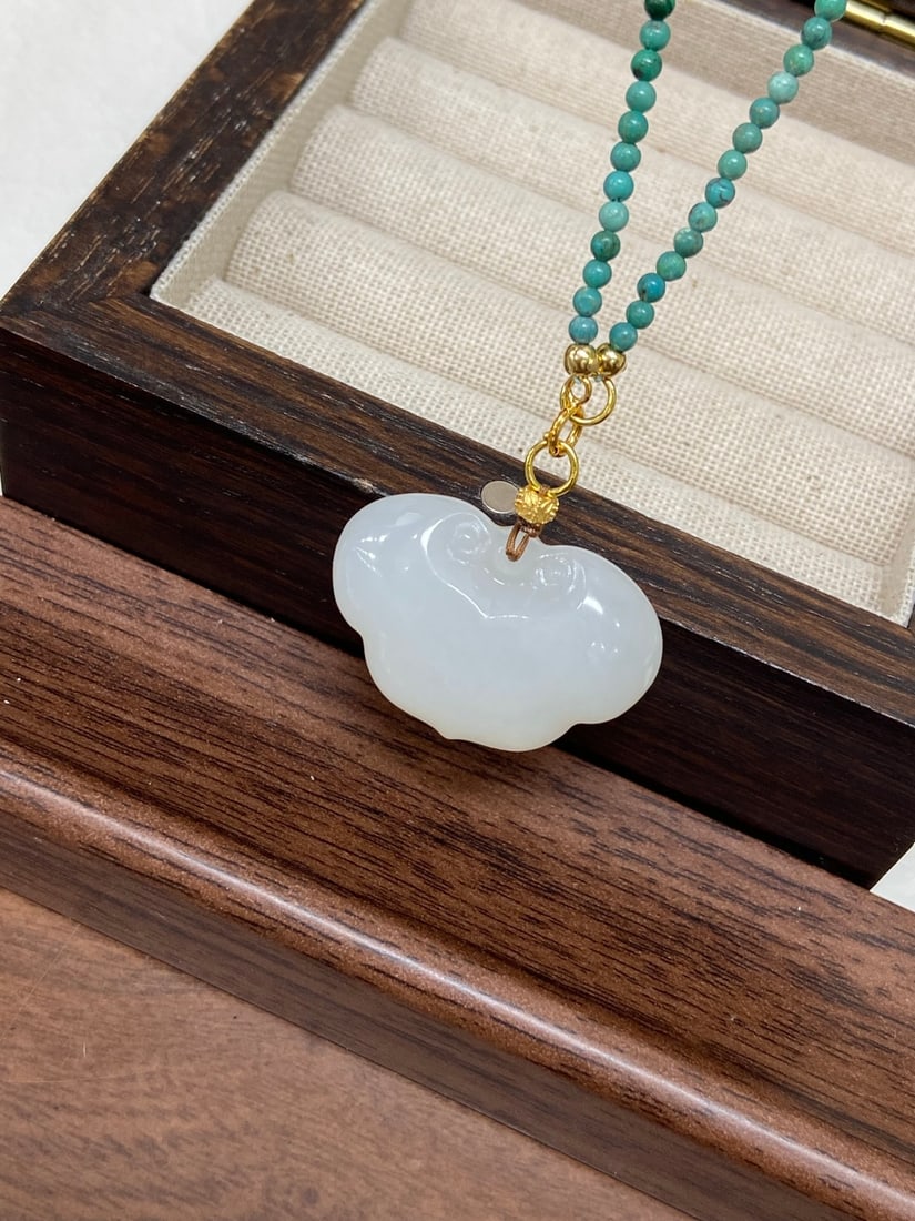 A Finely Carved Hetian Jade 'Noubo' White Jade 'Auspicious Lock' Pendant: A finely carved Hetian jade pendant of 'noubo' (glutinous-icy) white quality, carved as an auspicious lock with cloud motifs, symbolizing protection and blessing. The jade is of fine, warm texture wit