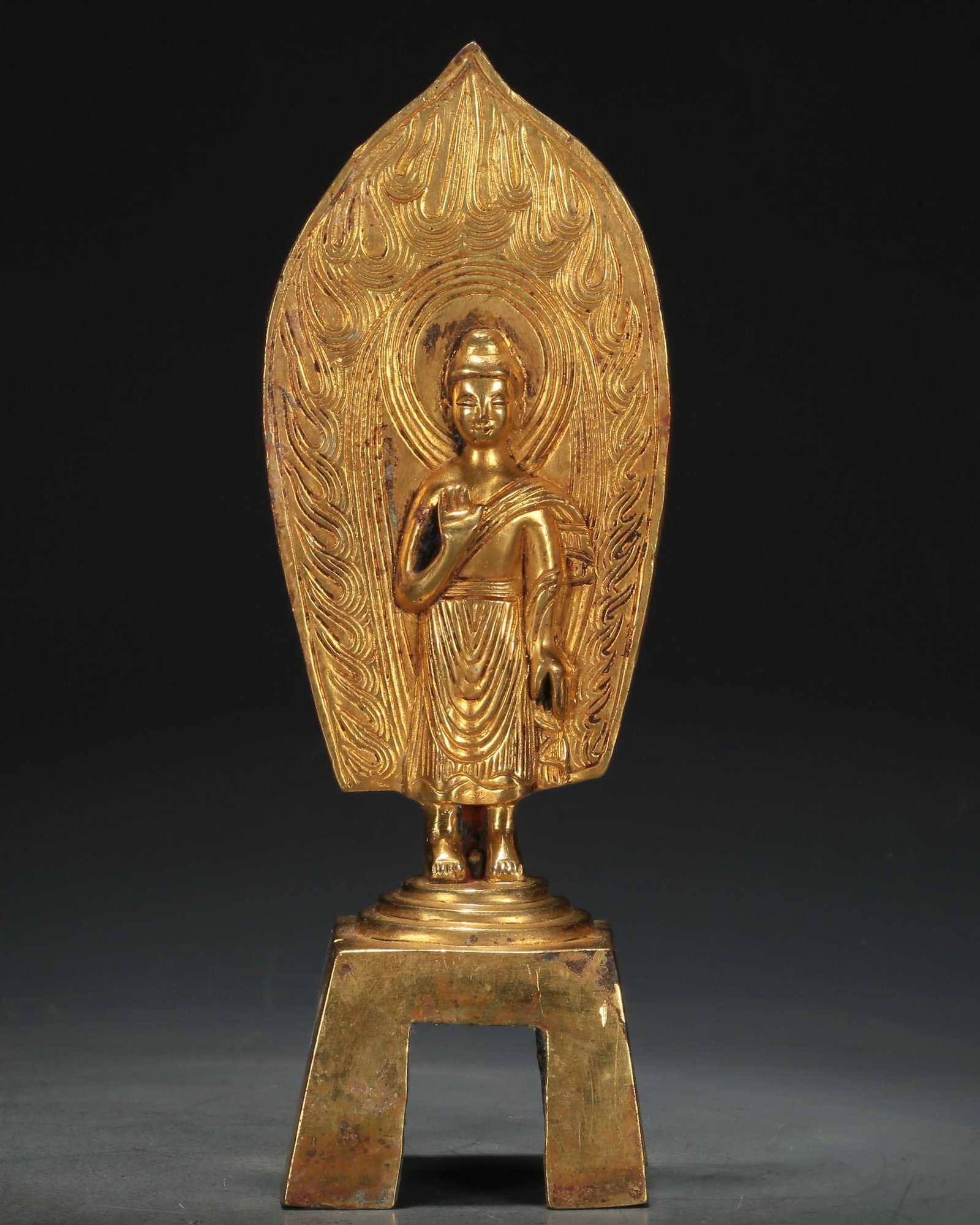 An Archaic Gilt-Bronze Figure Of Buddha Shakyamuni: An Archaic Gilt-Bronze Figure Of Buddha Shakyamuni. The figure is stood on a high base, hair tied into a high chignon, body rendered clad in long, flowing robes, the voluminous drapery elegantly casca