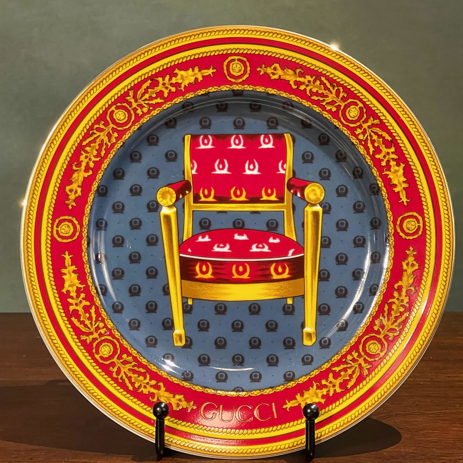 Gucci Porcelain Collectors Item Gilded Chair Plate #1: Porcelain dessert plate with historical Napoleonic style chair by Gucci features gilded edging, blue background with the Gucci name incorporated into the Vermillion red-colored border at the bottom, a