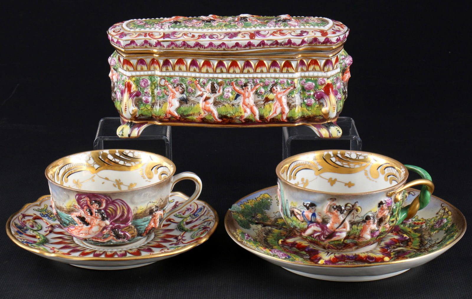 Capodimonte 2 tea cups and lidded box with cherub scene,: 5-piece porcelain, blue company mark, cherub scene in relief, polychrome painted, 2x tea cups H 5 cm x D 9 cm, 2x saucers D 15 - 17 cm, 1x lidded box H 8.5 cm x L 18 cm x W 7.5 cm, chip on one cup rim