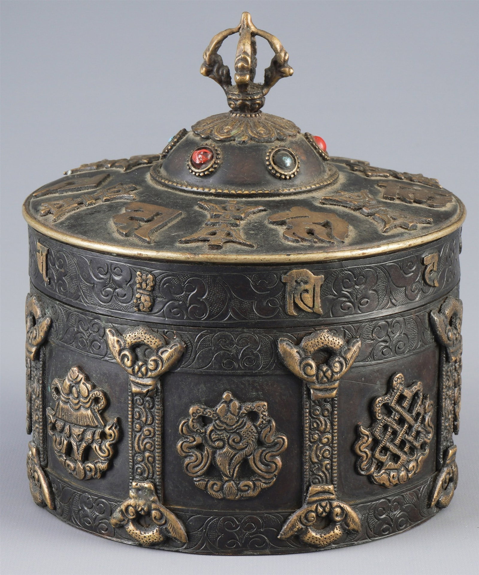 Bronze box with eight lucky symbols, Tibet, 20th century: Bronze, Tibet, 20th century, casket with eight lucky symbols, cylindrical shape with lid, the walls divided by column and tendril motifs, inscriptions in Tibetan script on the lid, H. 18 cm × D. 15