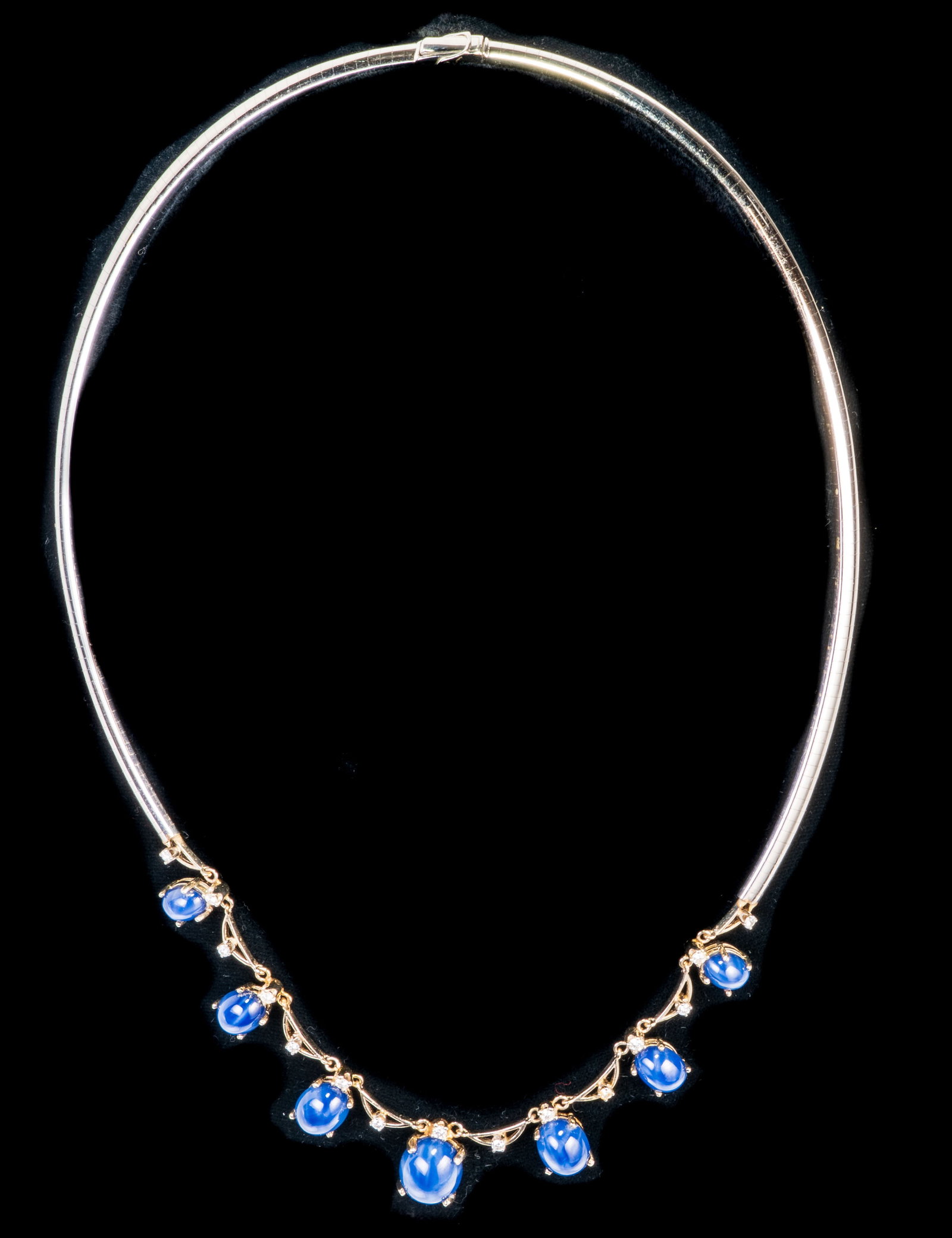 14k White Gold Synthetic Star Sapphire and Diamond Necklace: 14k white gold omega necklace with seven cabochon synthetic star sapphire pendants and 13 small round cut diamonds (0.01 ct. each). Measurements: The necklace is approximately 17 inches long with the 